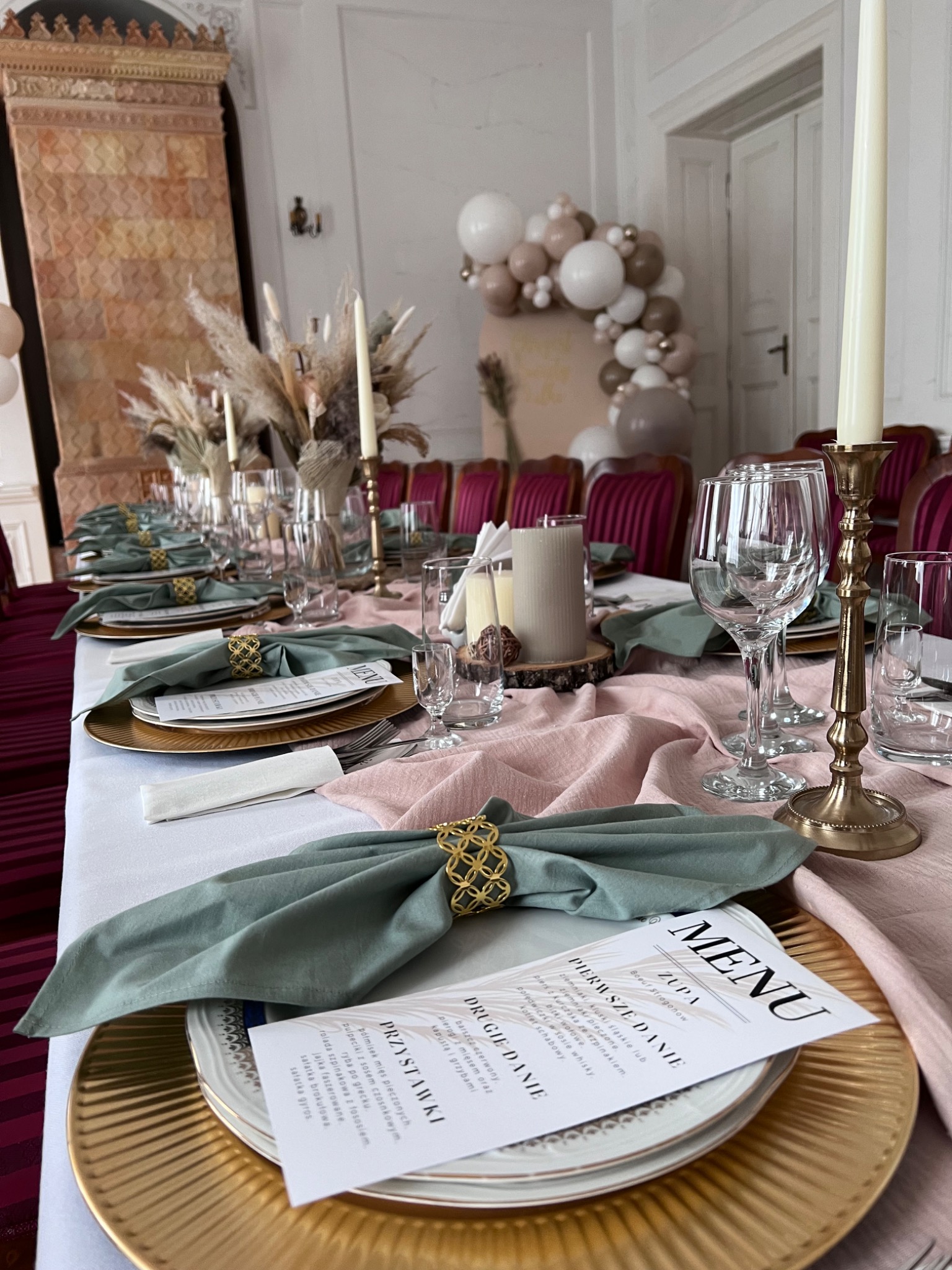 Uroczysta aranżacja stołu z elegancką zastawą, lnianymi serwetkami ze złotymi pierścieniami, menu, świecami i kompozycją z traw pampasowych, całość utrzymana w pastelowej kolorystyce z dekoracją...