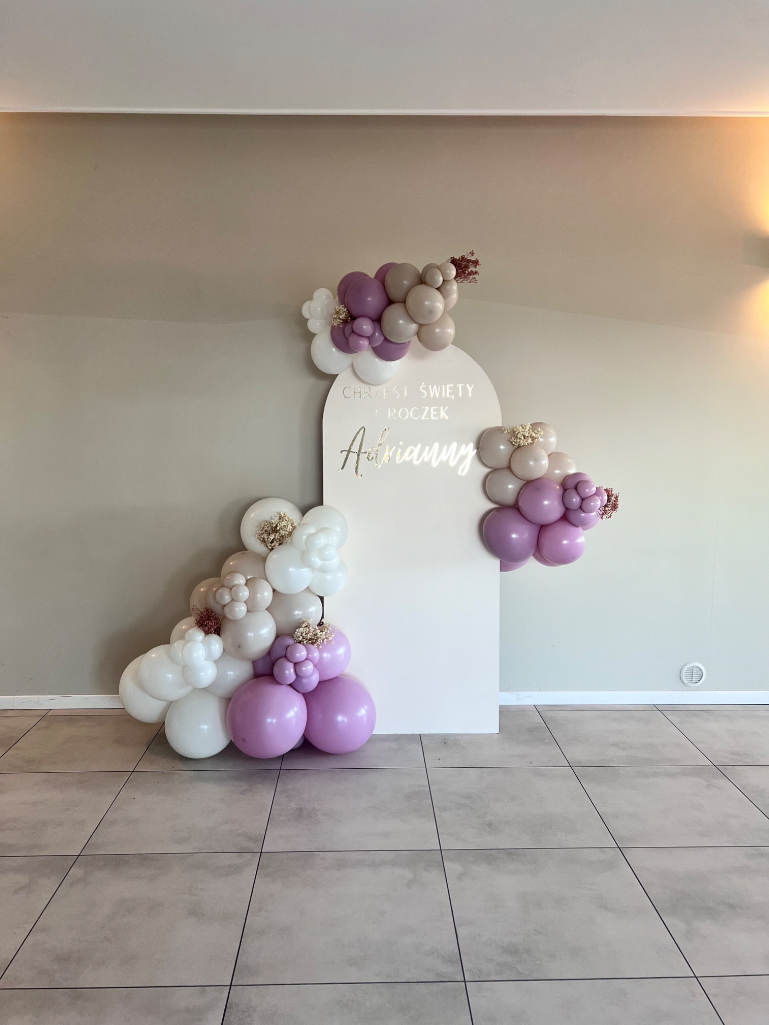Dekoracja z białych i fioletowych balonów oraz suszonych kwiatów na roczek dziecka, z napisem na białej tablicy.