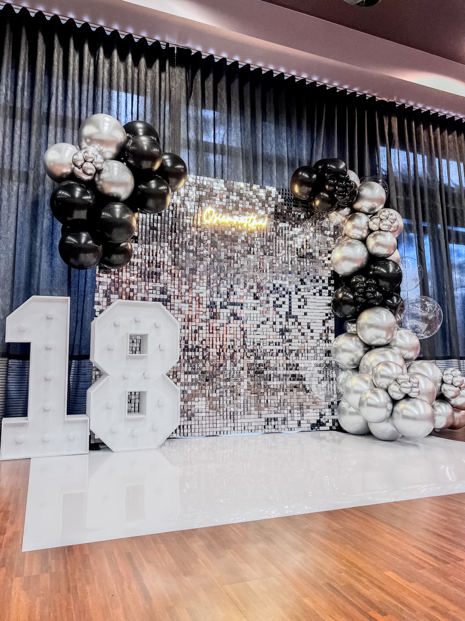 Dekoracja na osiemnaste urodziny: srebrna ściana z błyszczących kwadratów, neonowy napis, balony w kolorach srebrnym i czarnym oraz podświetlane cyfry 18.