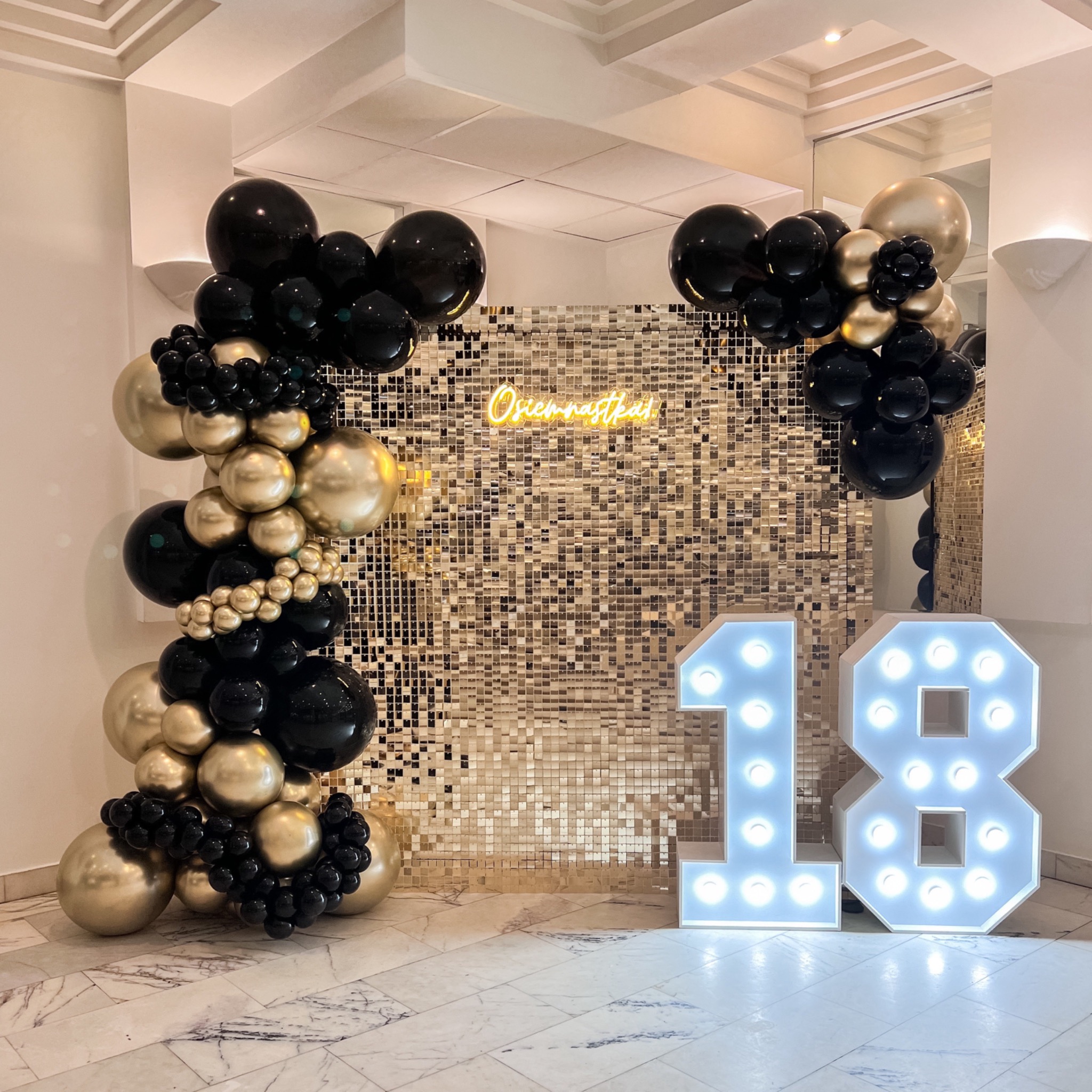 Dekoracja na osiemnaste urodziny: złota ściana z cekinów, neonowy napis 'Osiemnastka!' oraz balonowa girlanda w kolorach czerni i złota, obok podświetlane cyfry '18'.