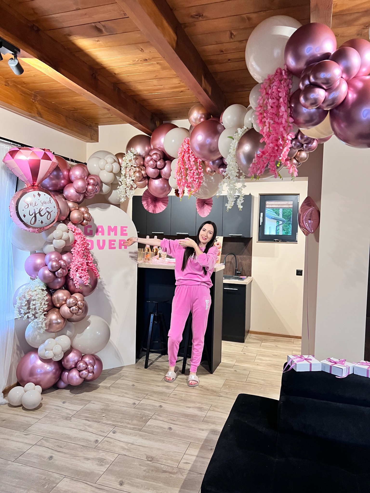 Dekoracja balonowa w odcieniach różu i bieli z napisem 'Game Over' i balonem w kształcie diamentu 'She Said Yes', kobieta w różowym dresie pozuje na tle kuchni.