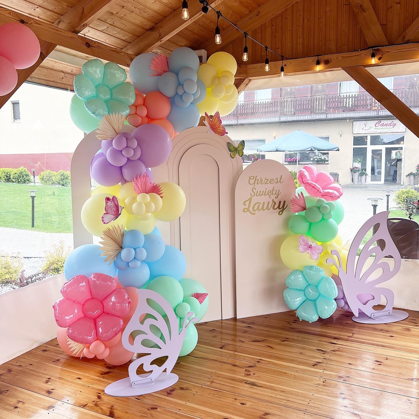 Dekoracja balonowa w pastelowych kolorach na Chrzest Święty Laury, z motywem motyli, na drewnianym tarasie restauracji Candy.