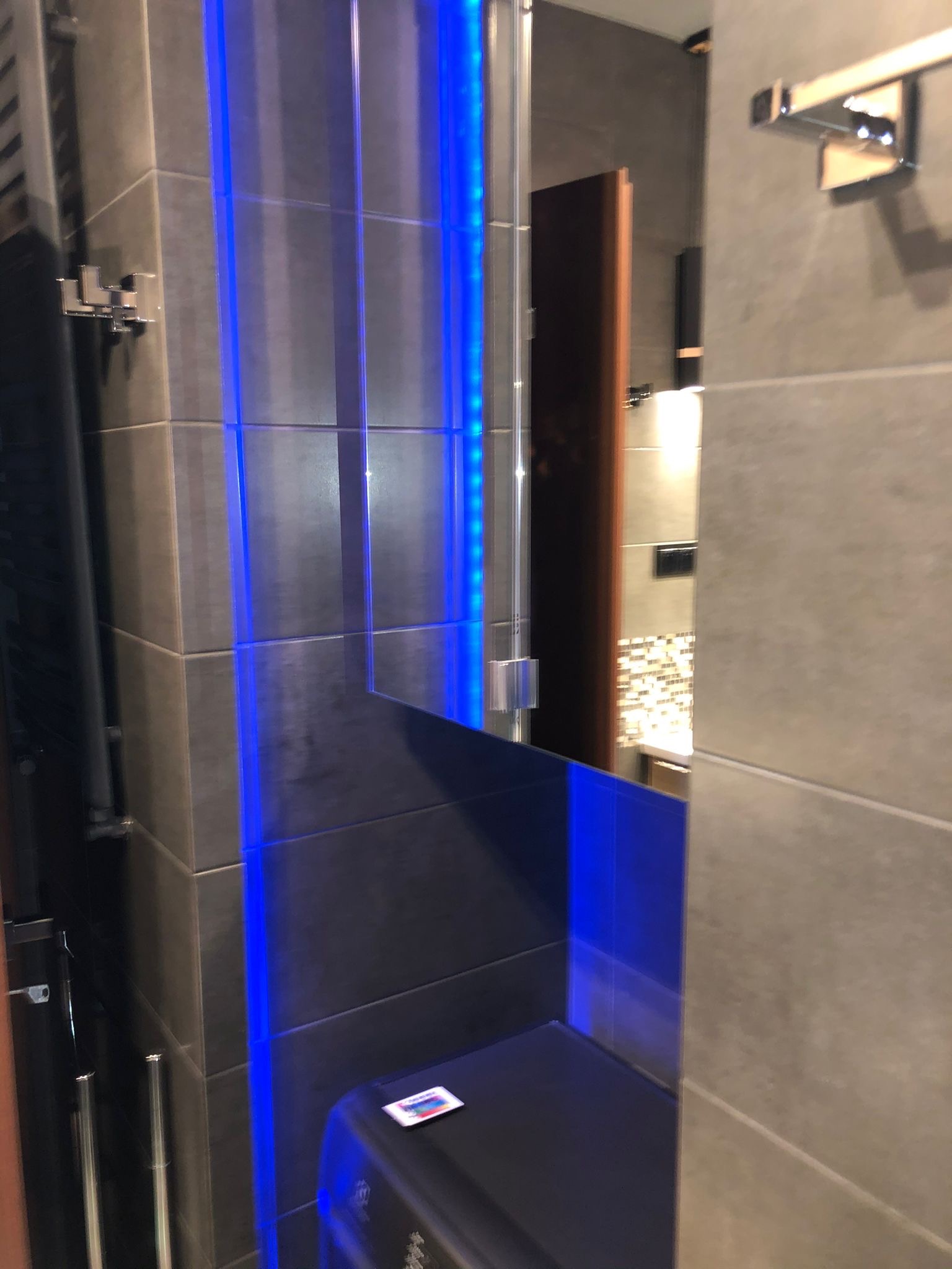 Fragment łazienki z szarymi płytkami, oświetleniem LED w kolorze niebieskim wzdłuż kabiny prysznicowej oraz widocznym fragmentem umywalki z mozaiką w lustrze.