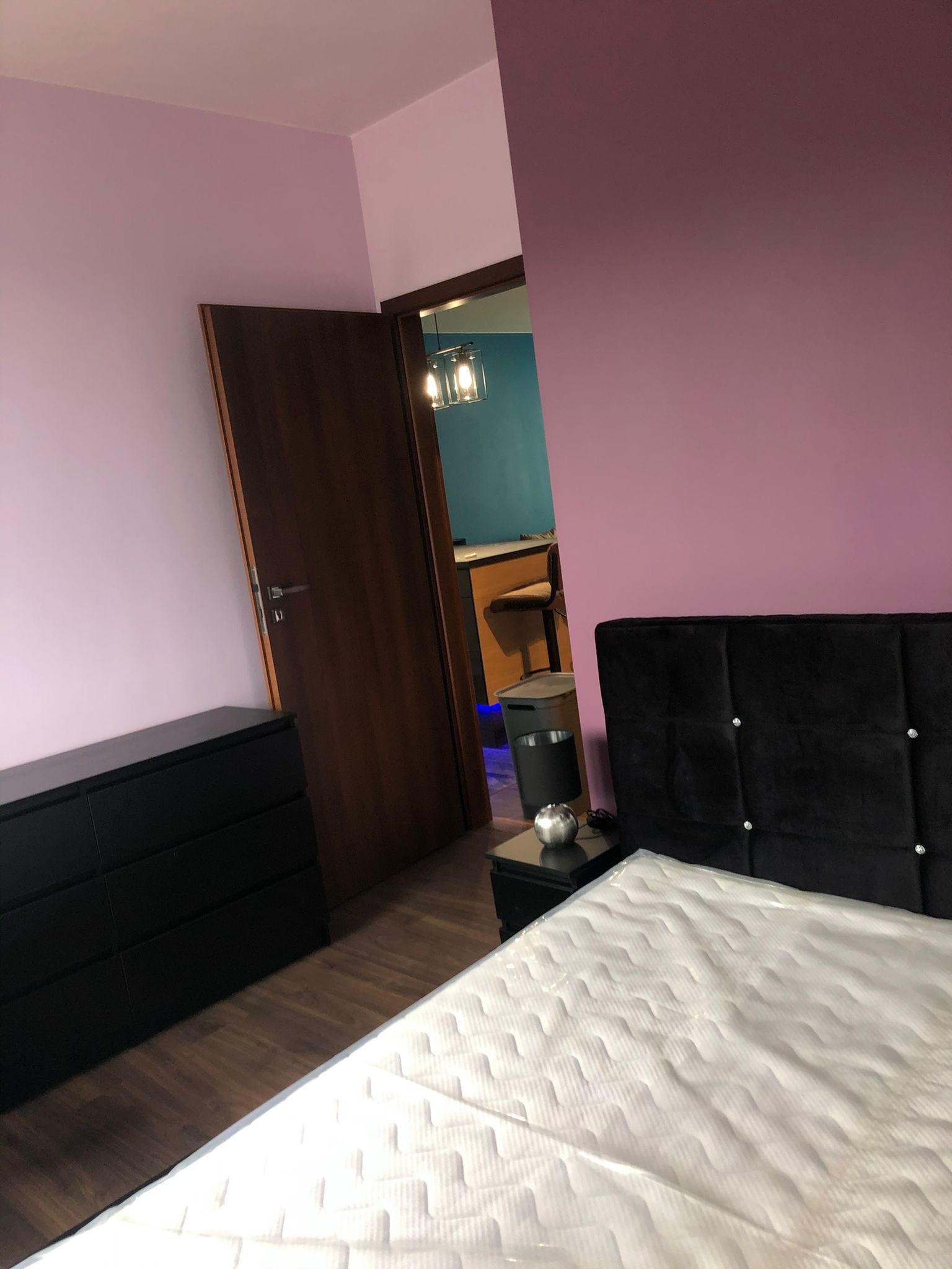 Sypialnia z łóżkiem z czarnym wezgłowiem, fioletowe ściany, drewniana podłoga i czarna komoda, widok na pokój z niebieską ścianą przez otwarte drzwi.