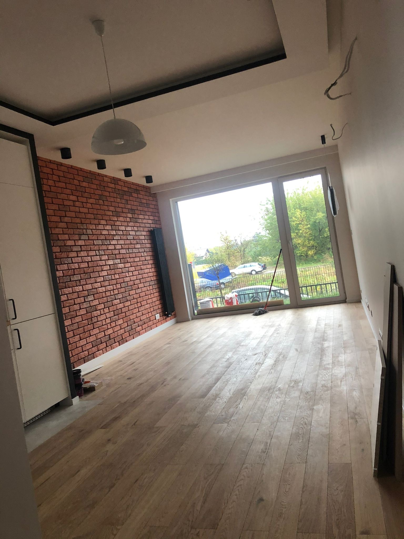 Pokój po remoncie z ceglaną ścianą, drewnianą podłogą i dużym oknem z widokiem na zewnątrz, widoczne niedokończone prace przy suficie.