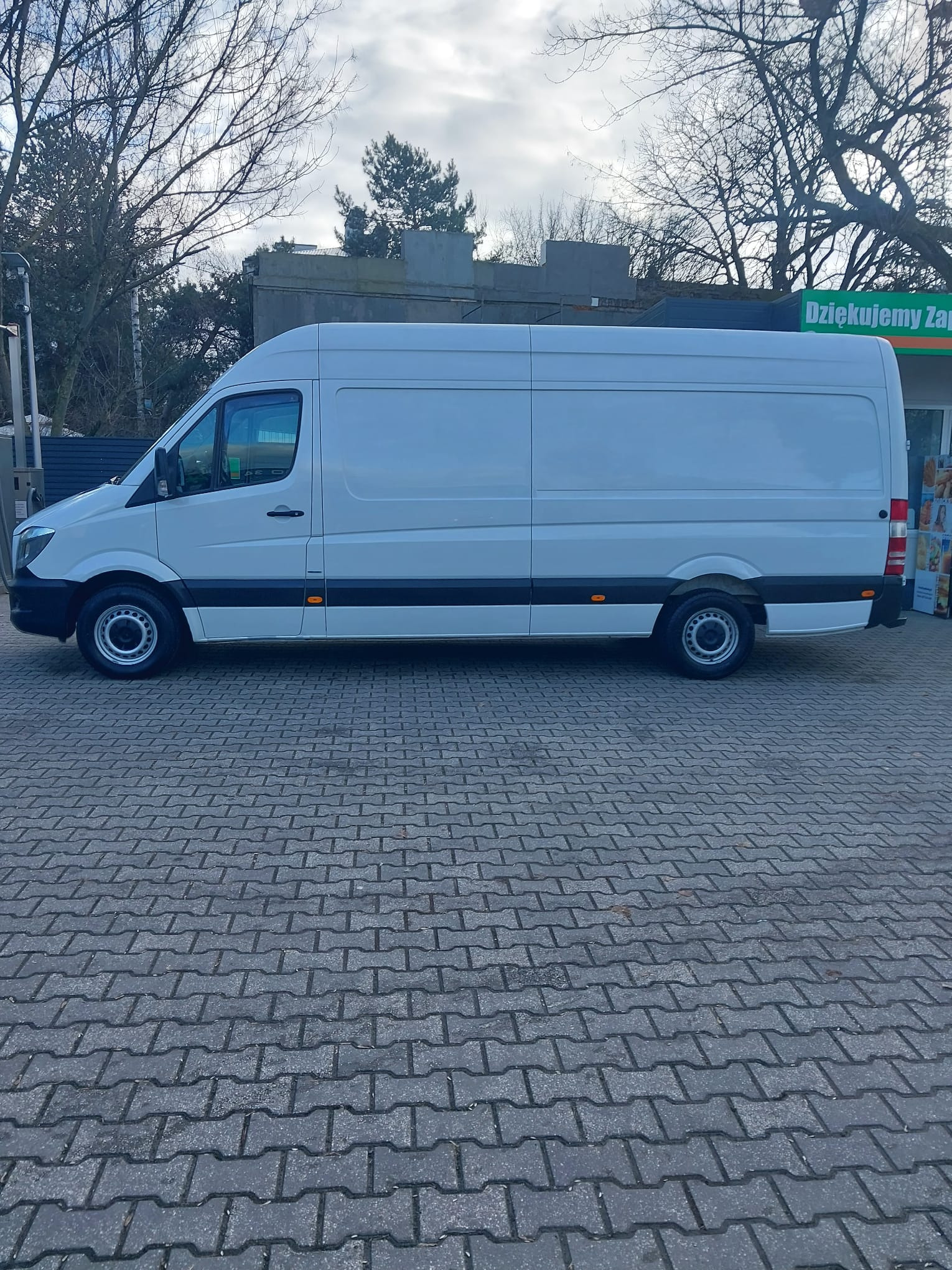 Biały, długi bus dostawczy Mercedes-Benz Sprinter zaparkowany na brukowanym parkingu w pochmurny dzień, widok z boku.