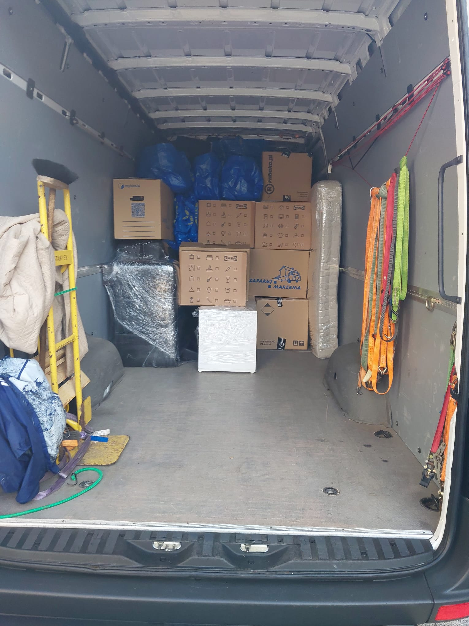 Wnętrze busa załadowane kartonami, meblami owiniętymi folią i zabezpieczone pasami transportowymi, gotowe do transportu. Widoczna drabina i ubrania robocze.