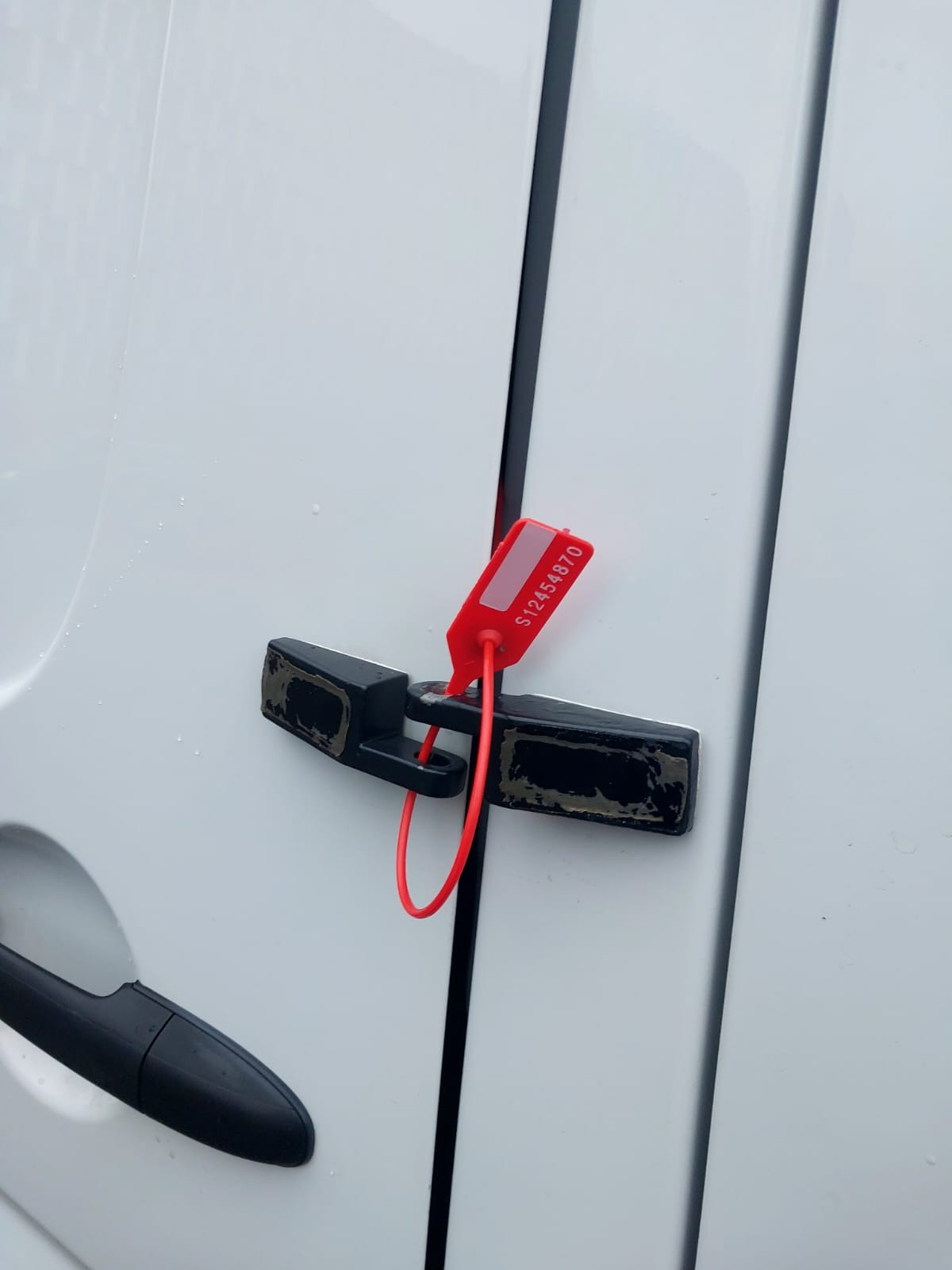 Zamknięte drzwi białego busa z czarnym zamkiem zabezpieczonym czerwoną plombą z numerem seryjnym S12454870.