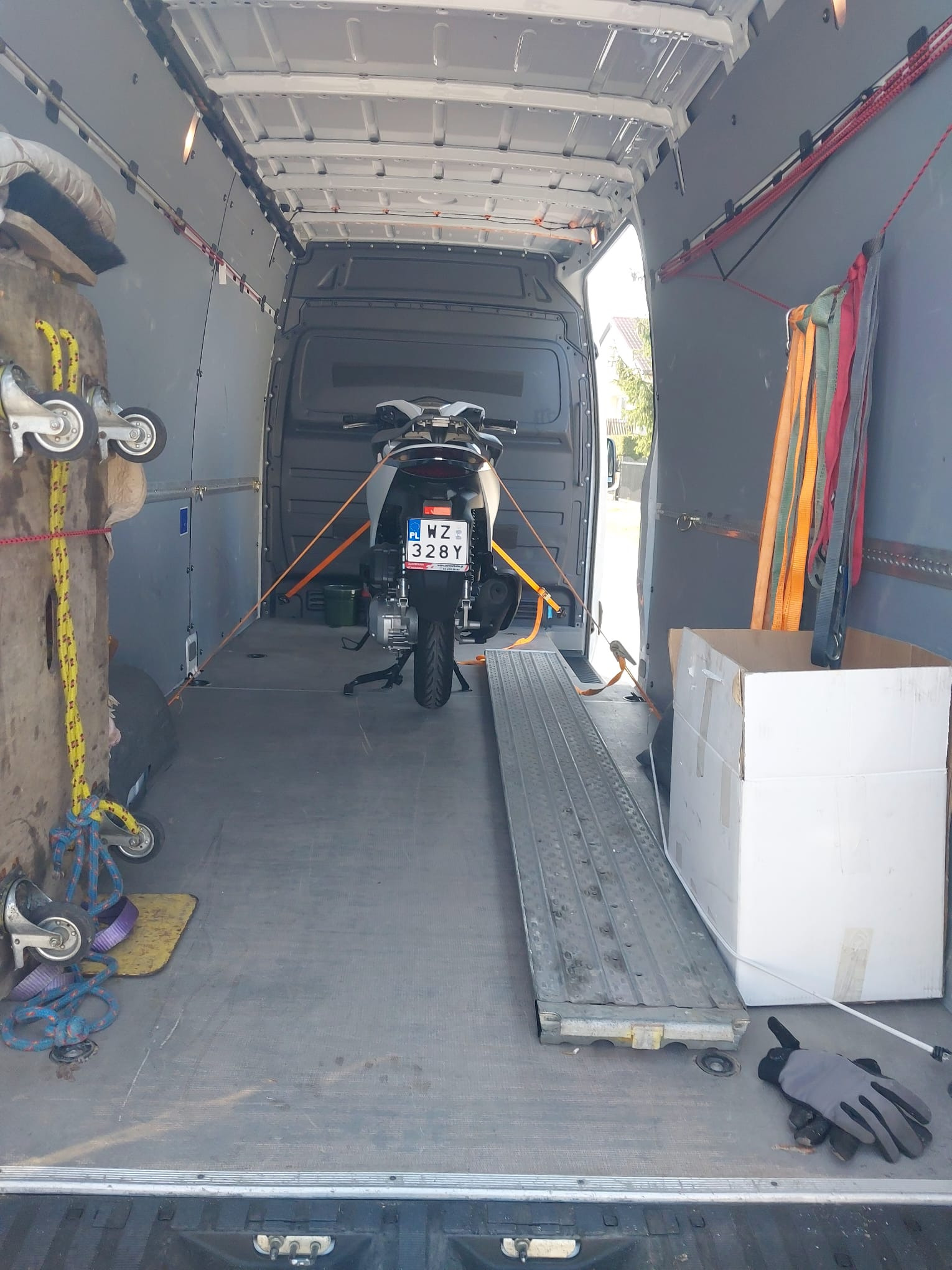 Wnętrze busa: motocykl zabezpieczony pasami transportowymi, najazd, karton, wózek transportowy z kółkami, rękawice robocze.
