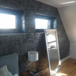 Pokój na poddaszu z tapetą w motywy miejskie, drewnianą podłogą, lustrem stojącym i szafką nocną z lampką.