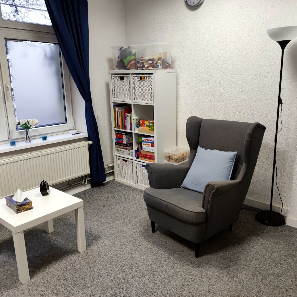 Gabinet z szarym fotelem typu uszak, białym regałem z książkami i zabawkami, oraz stolikiem z chusteczkami i zegarem, oświetlony lampą stojącą.
