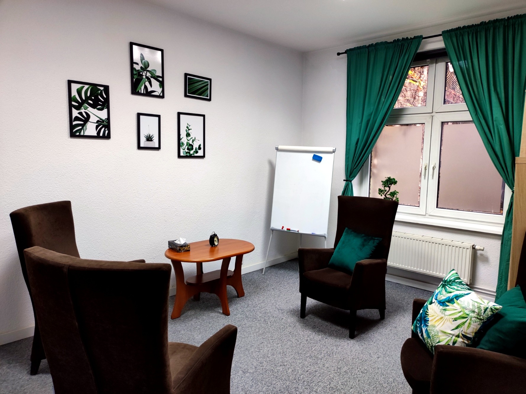 Jasny gabinet z brązowymi fotelami, drewnianym stolikiem z chusteczkami i zegarem, tablicą suchościeralną, zielonymi zasłonami i dekoracjami roślinnymi na ścianie.