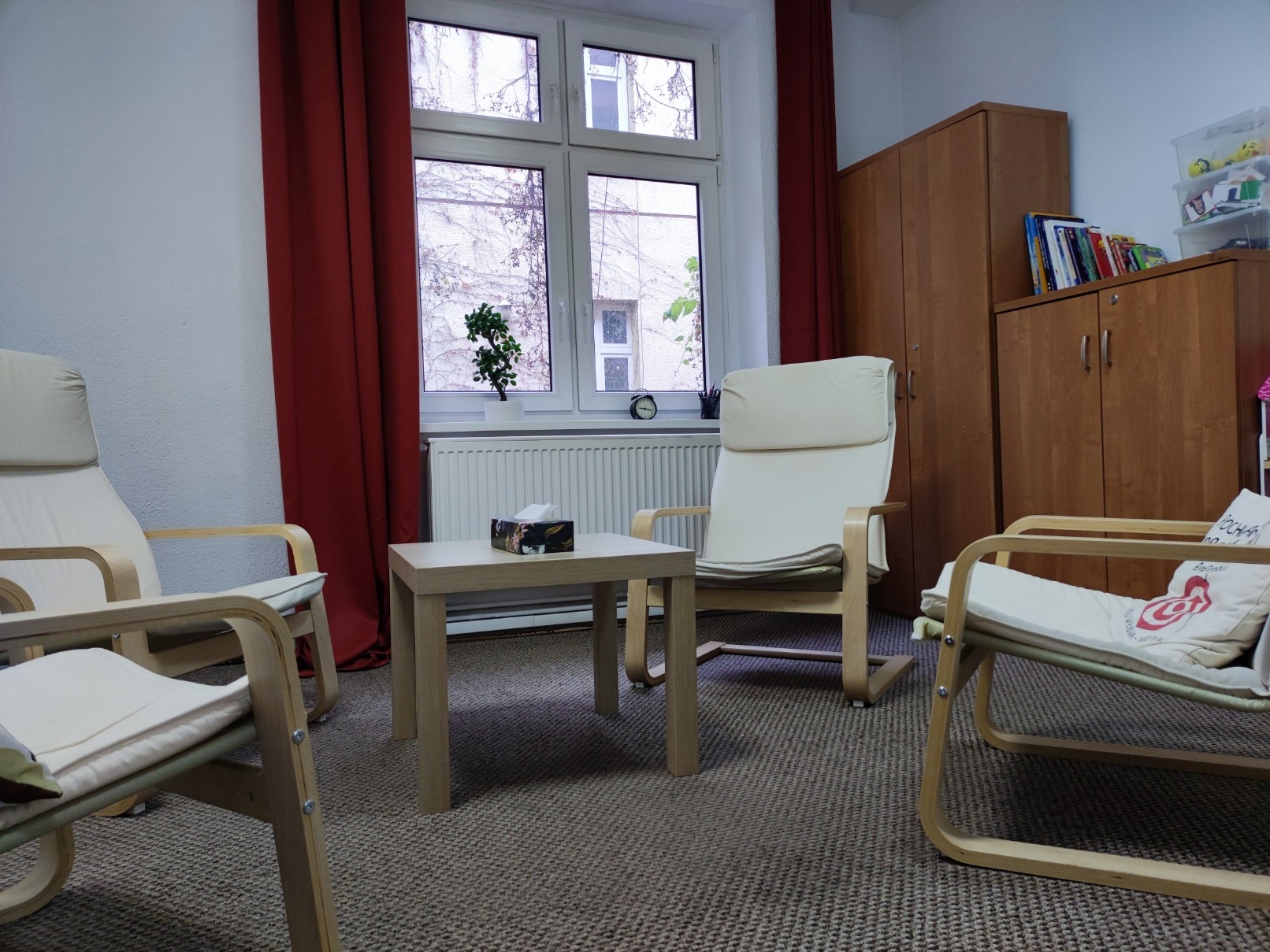 Jasne, minimalistyczne wnętrze gabinetu z trzema drewnianymi fotelami z kremowymi poduszkami, małym stolikiem z pudełkiem chusteczek, brązową szafą i oknem z widokiem na budynek porośnięty bluszczem.