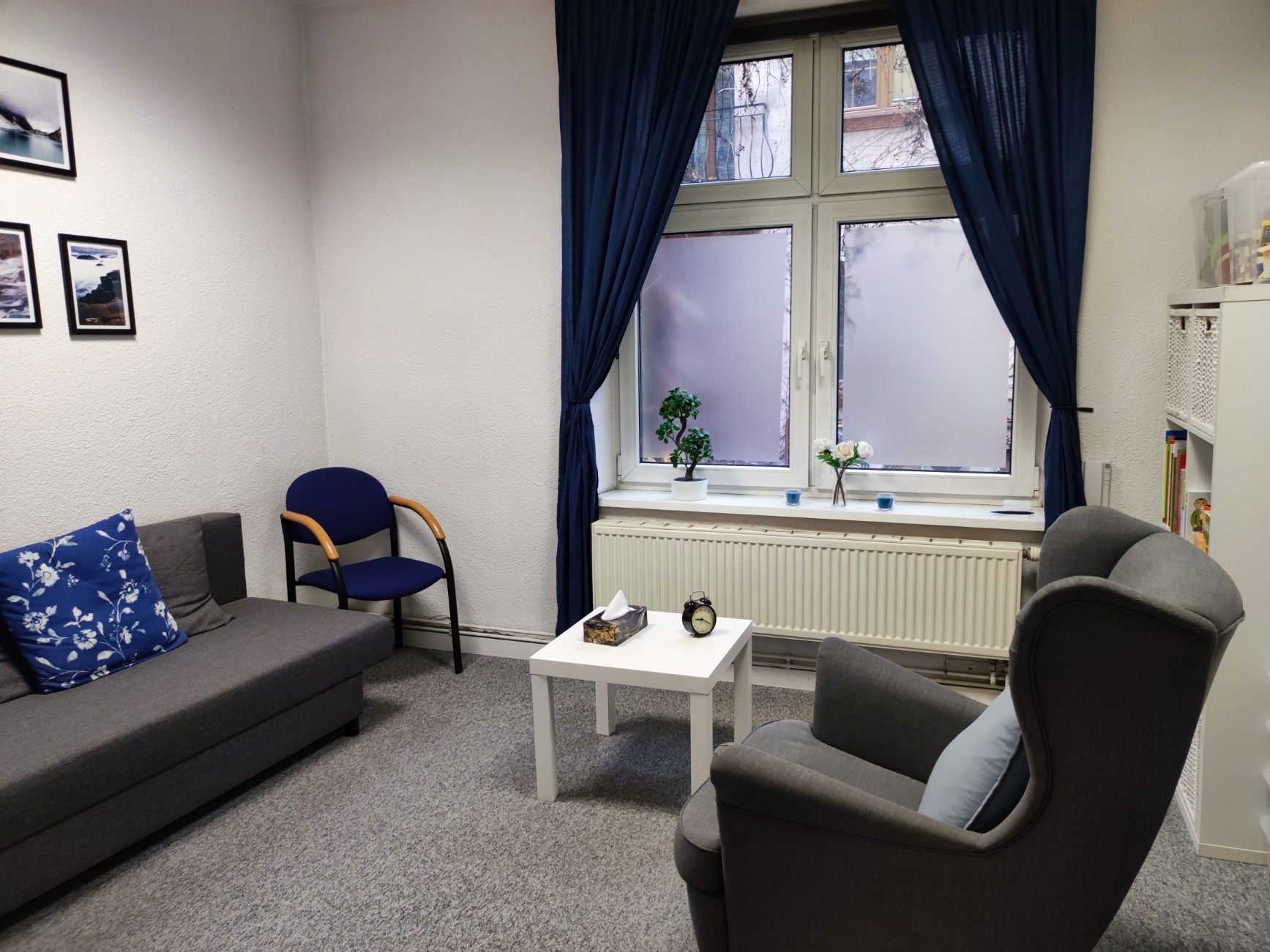 Jasny pokój z szarym dywanem, kanapą z poduszką w kwiaty, krzesłami, białym stolikiem z chusteczkami i zegarem, oraz oknem z niebieskimi zasłonami i roślinami na parapecie.