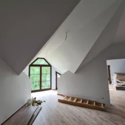 Poddasze po remoncie z drewnianą podłogą, jasnymi ścianami i oknem szczytowym. Na podłodze leżą panele podłogowe i drabina, widoczne też środki czystości.
