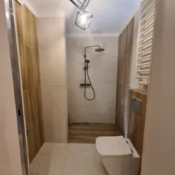 Nowoczesna łazienka z kabiną prysznicową typu walk-in, wykończona płytkami imitującymi drewno i marmur, oświetlona geometryczną lampą wiszącą.