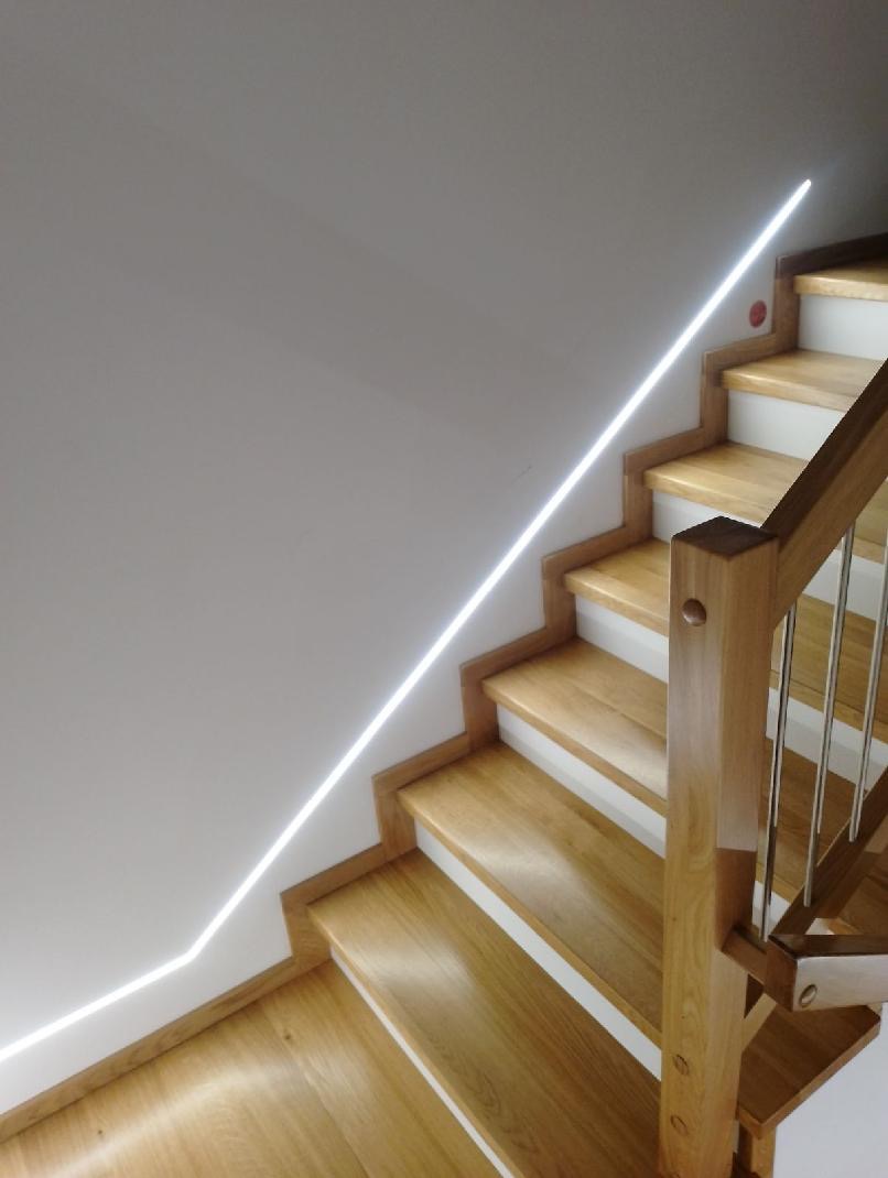 Schody drewniane z oświetleniem LED wzdłuż ściany, jasne drewno, białe podstopnice, metalowe barierki, widok z góry.