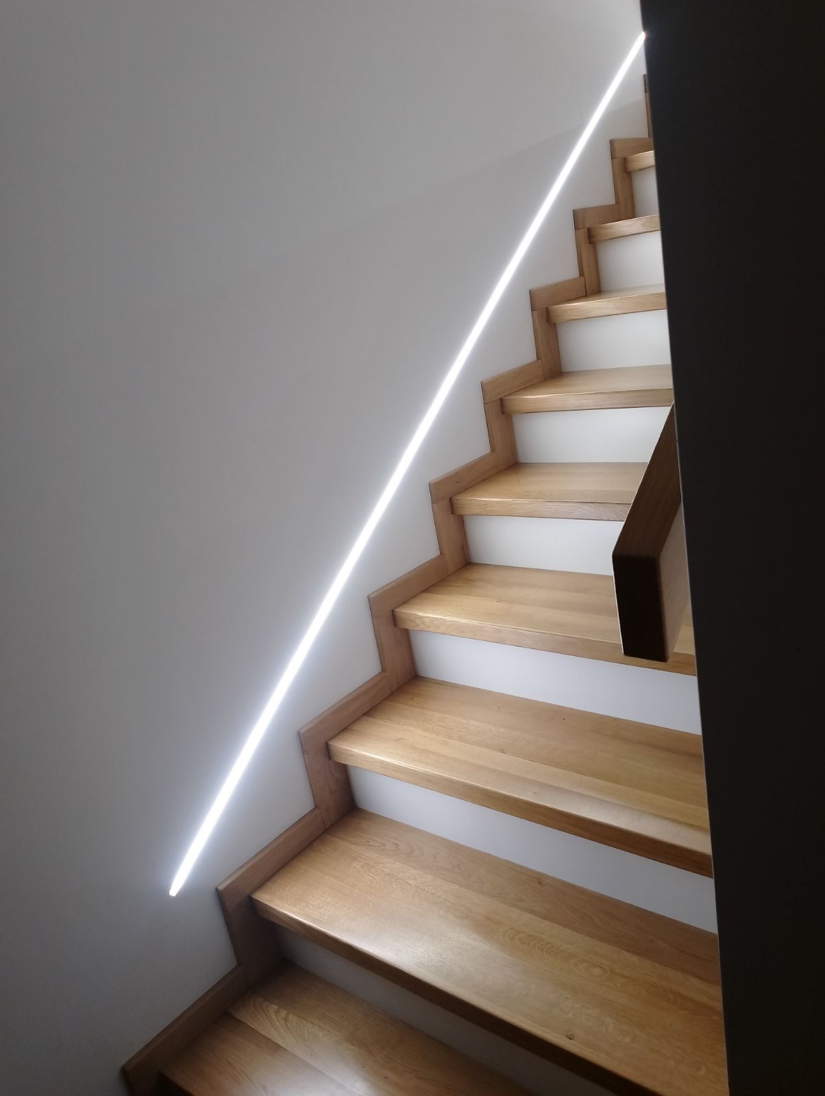 Oświetlenie LED schodowe wzdłuż ściany, drewniane stopnie z białymi podstopnicami, nowoczesny design, pionowa linia światła.