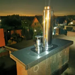 Błyszczący, metalowy komin na dachu budynku mieszkalnego, widok nocą na okoliczne domy i drzewa, oświetlone światłami.