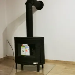 Czarny piec wolnostojący marki Invicta z podłączeniem do komina, stojący na szklanej płycie ochronnej, z widocznymi etykietami energetycznymi.