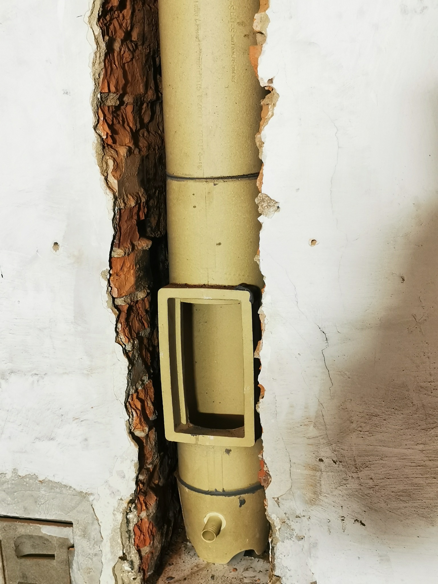 Żółta rura wentylacyjna z prostokątnym wlotem, zamontowana w bruździe w ścianie z odsłoniętą czerwoną cegłą i resztkami białego tynku.