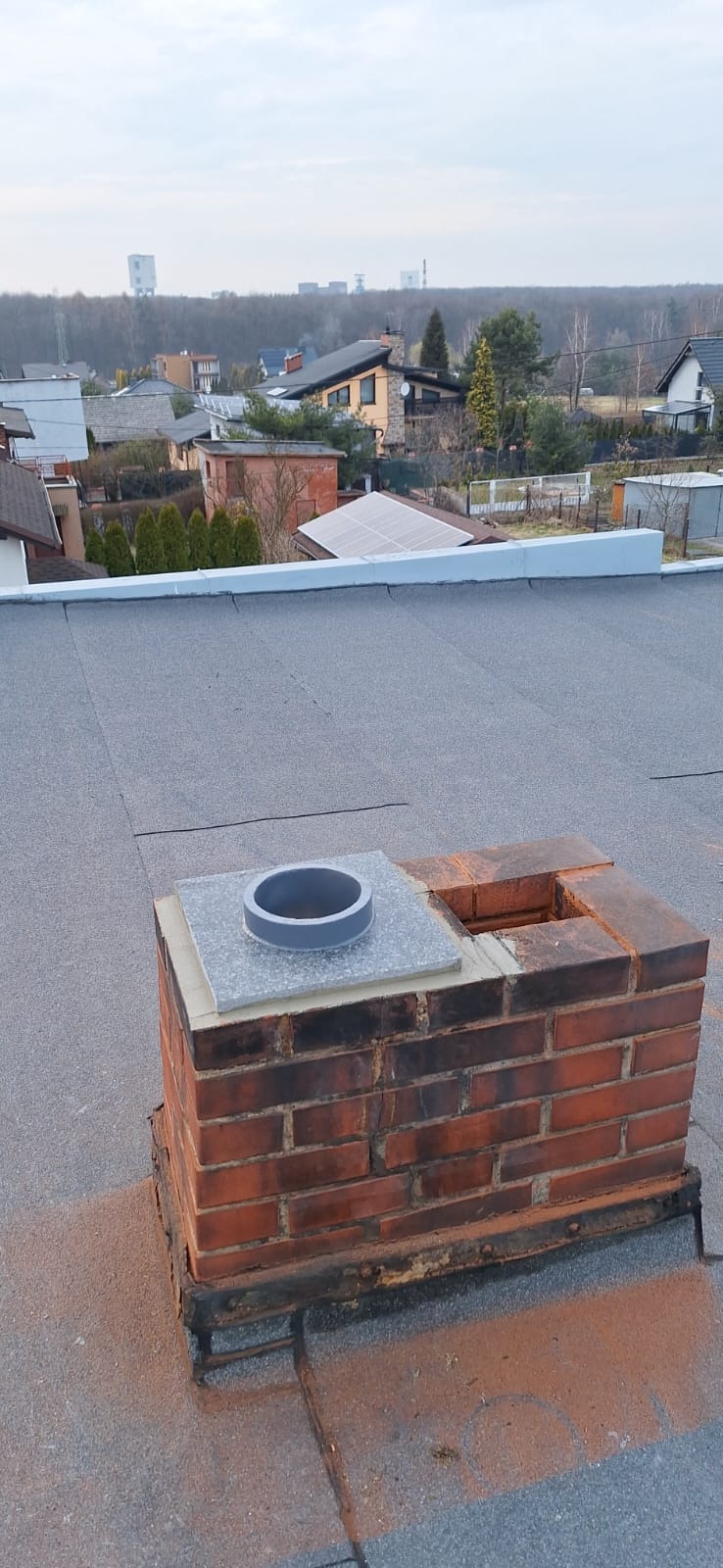 Ceglany komin na dachu z szarym wykończeniem i wkładem ceramicznym, widok z góry na tle zabudowań i drzew.