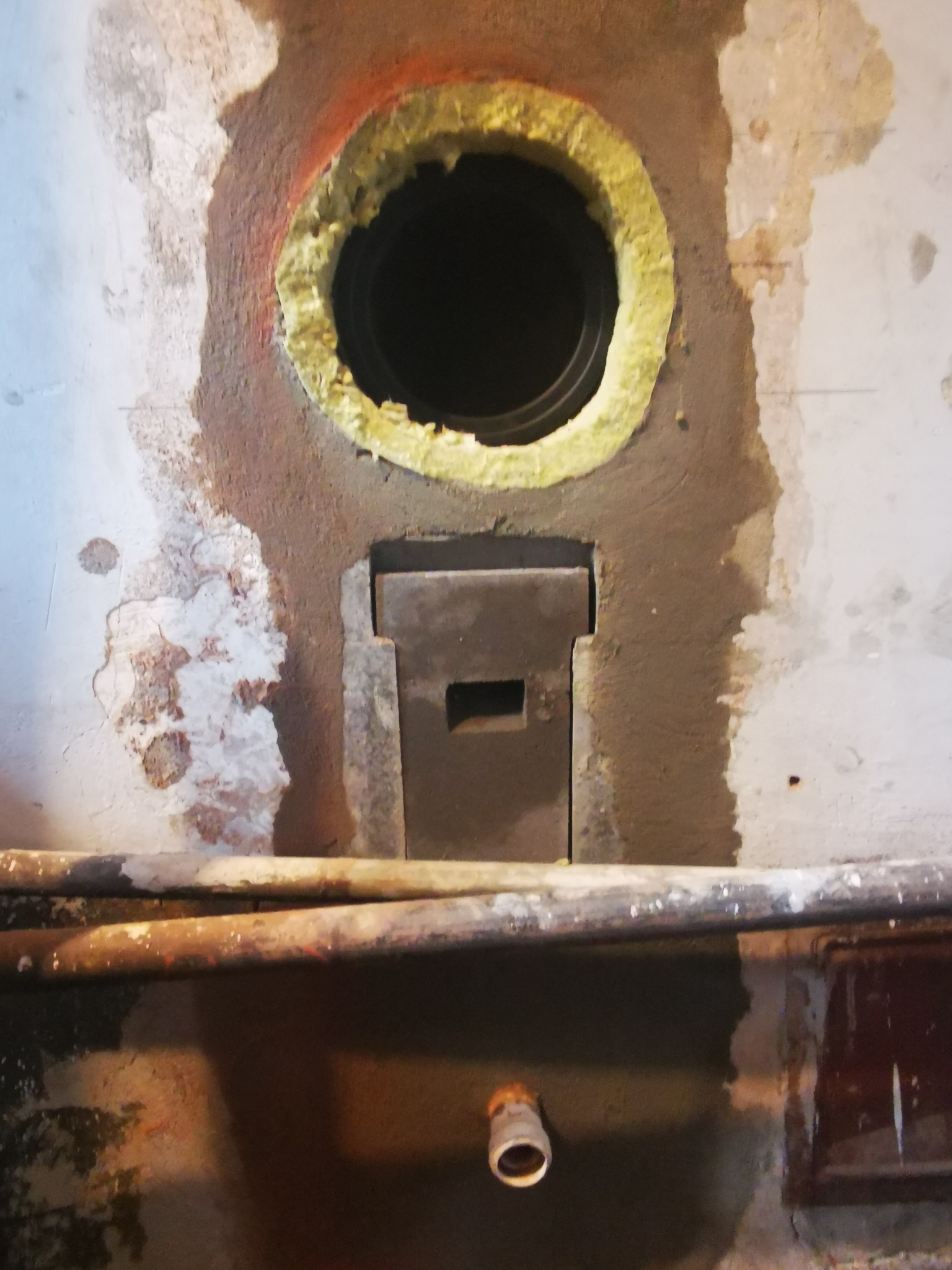 Fragment ściany z otworem wentylacyjnym obudowanym izolacją oraz metalową klapą rewizyjną, widoczne rury instalacyjne i ślady po remoncie.