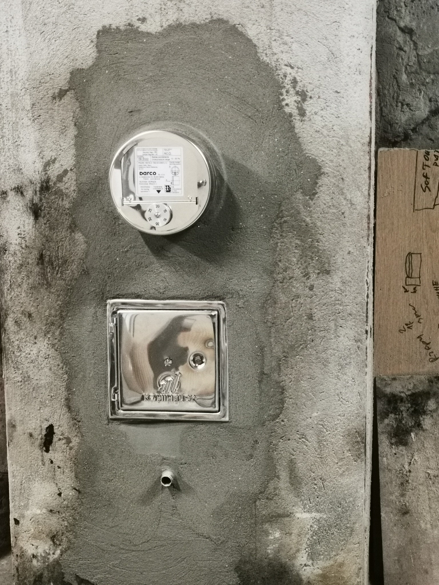 Świeżo zamontowany, chromowany licznik gazu i rewizja kominowa na szarej, otynkowanej ścianie, obok deska z odręcznym szkicem.