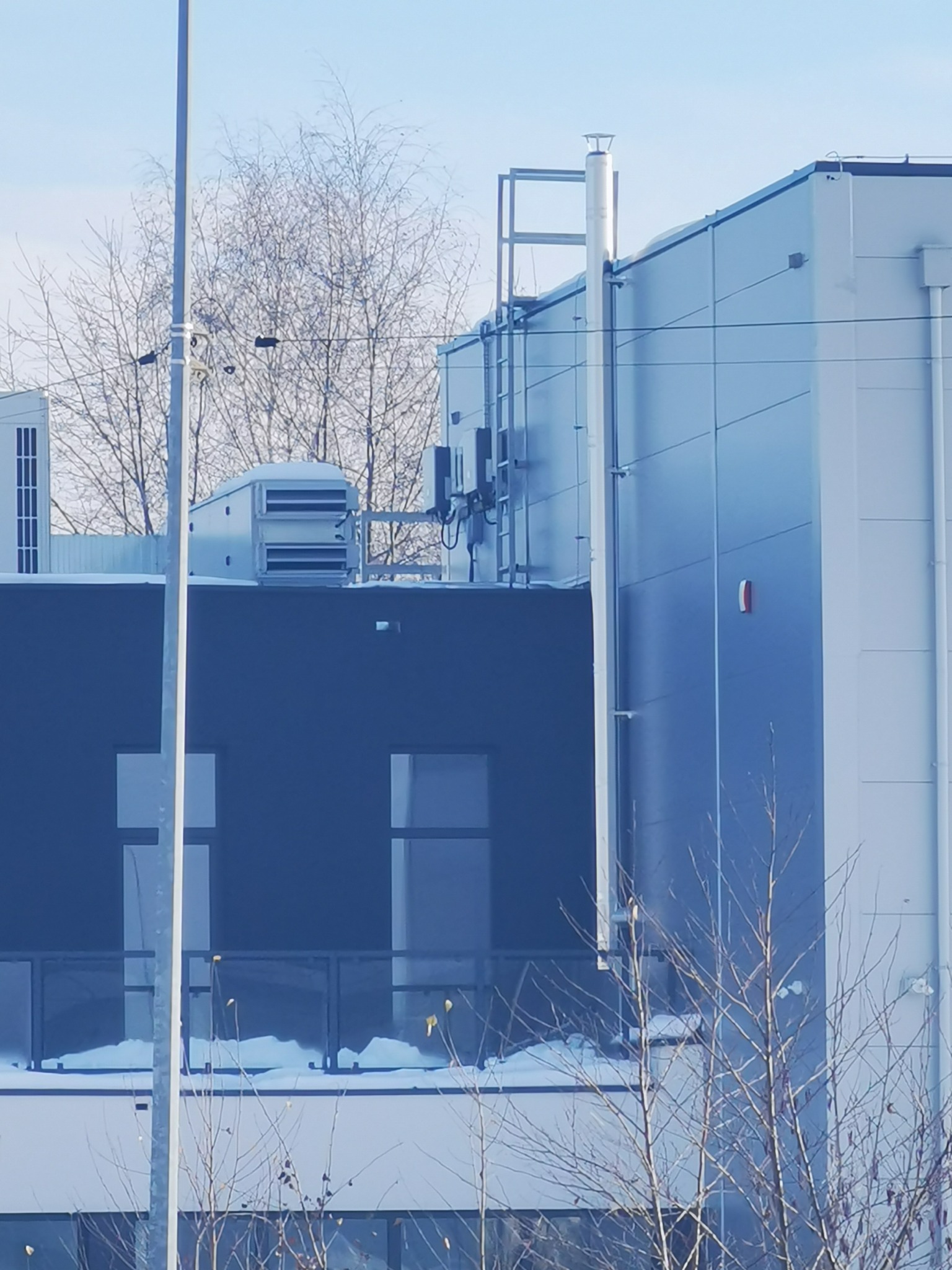 Zewnętrzny widok nowoczesnego budynku przemysłowego z widocznymi elementami wentylacji na dachu i kominem, uchwycony w zimowej scenerii.