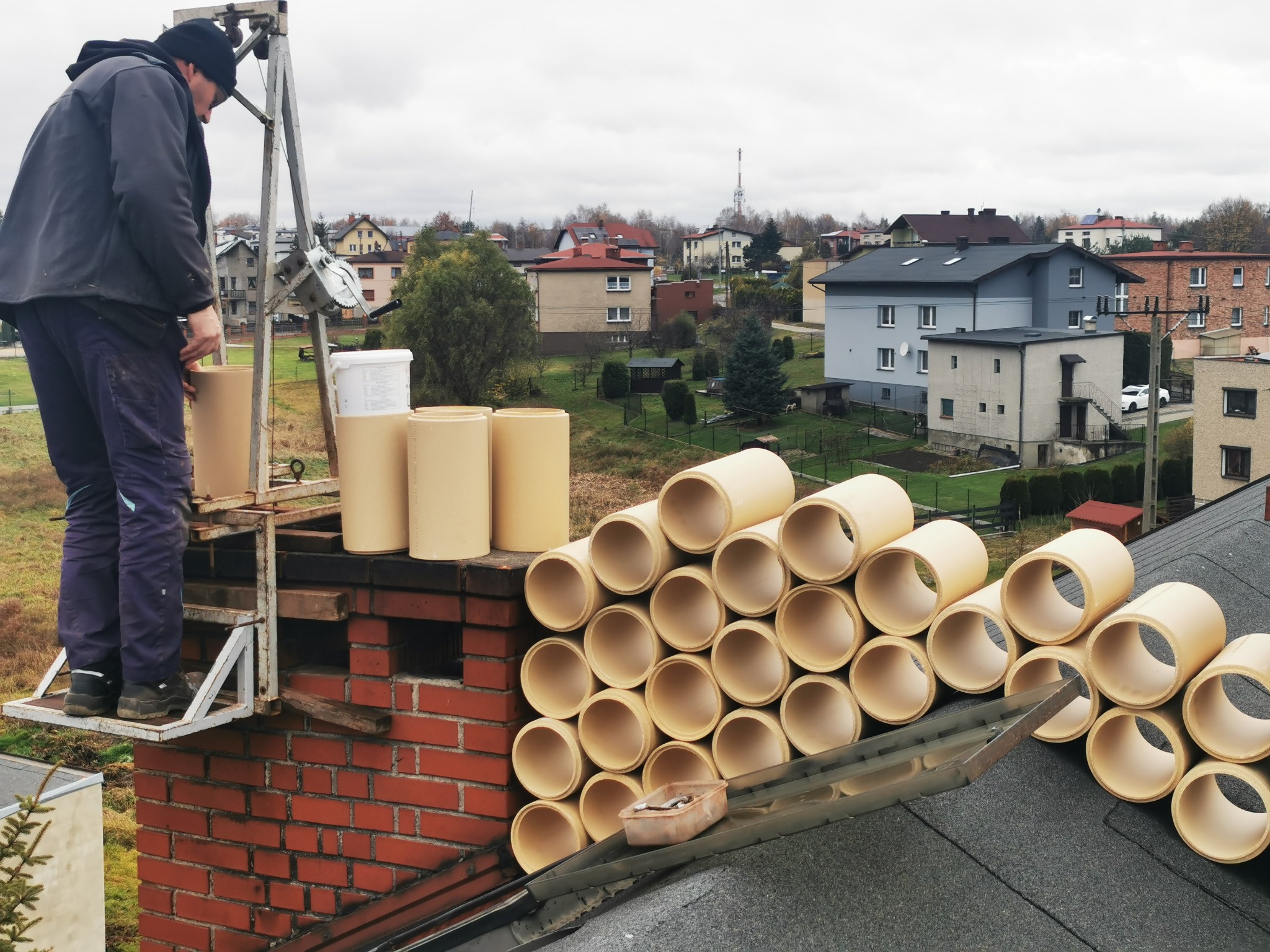 Kominiarz na rusztowaniu przy kominie z ułożonymi rurami ceramicznymi, przygotowujący się do montażu wkładu kominowego, widok z dachu.