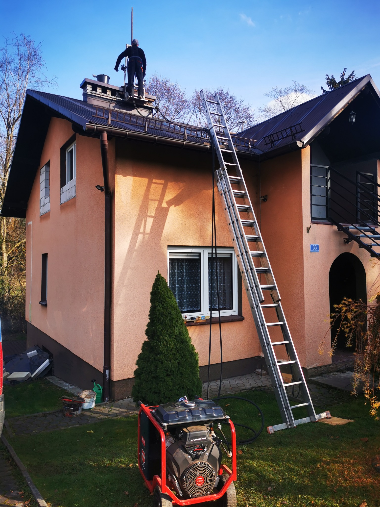 Pracownik w kombinezonie na dachu domu jednorodzinnego przy kominie, drabina oparta o ścianę, generator prądu na trawniku, błękitne niebo.