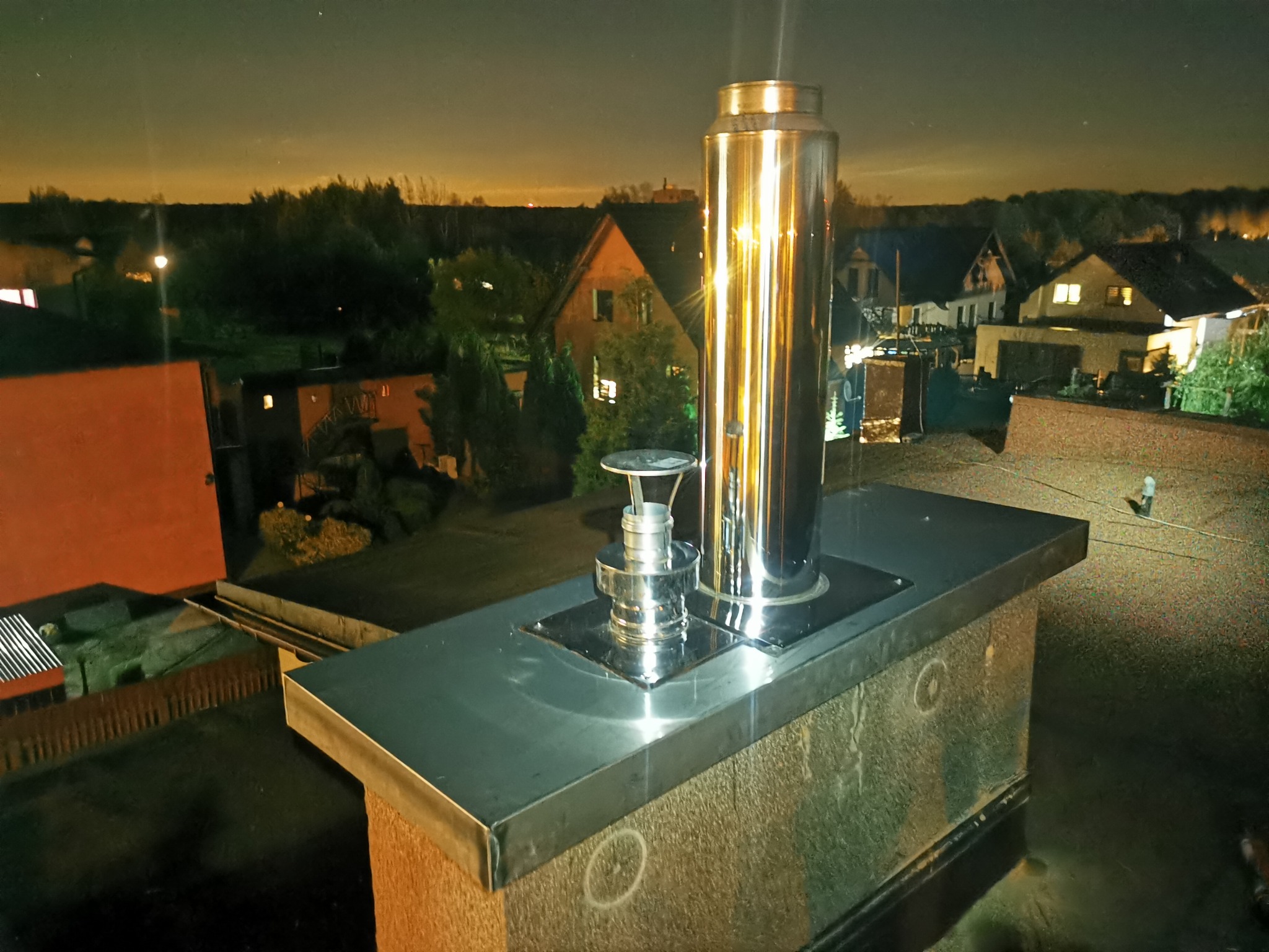 Błyszczący, metalowy komin na dachu budynku mieszkalnego, widok nocą na okoliczne domy i drzewa, oświetlone światłami.