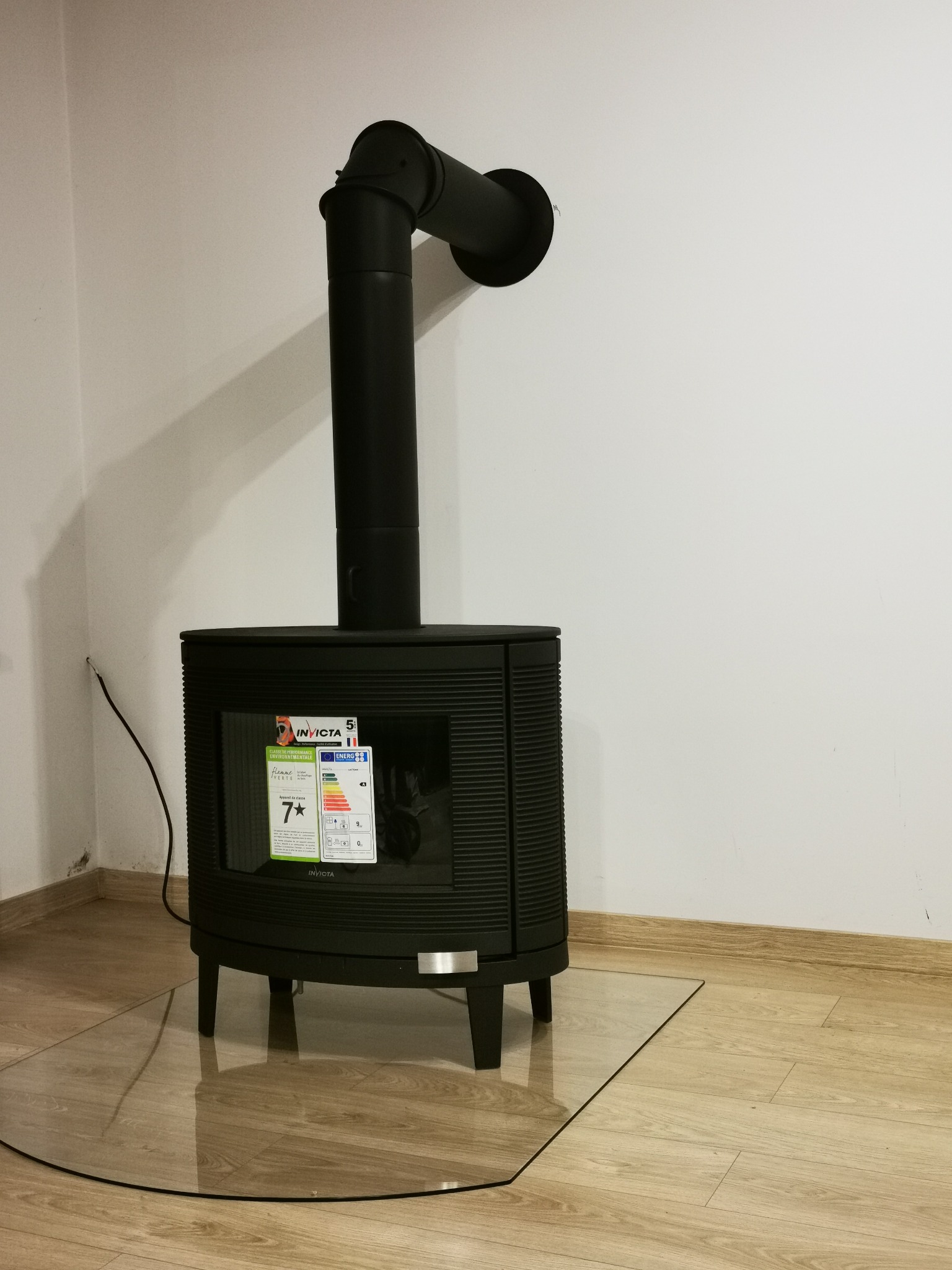 Czarny piec wolnostojący marki Invicta z podłączeniem do komina, stojący na szklanej płycie ochronnej, z widocznymi etykietami energetycznymi.