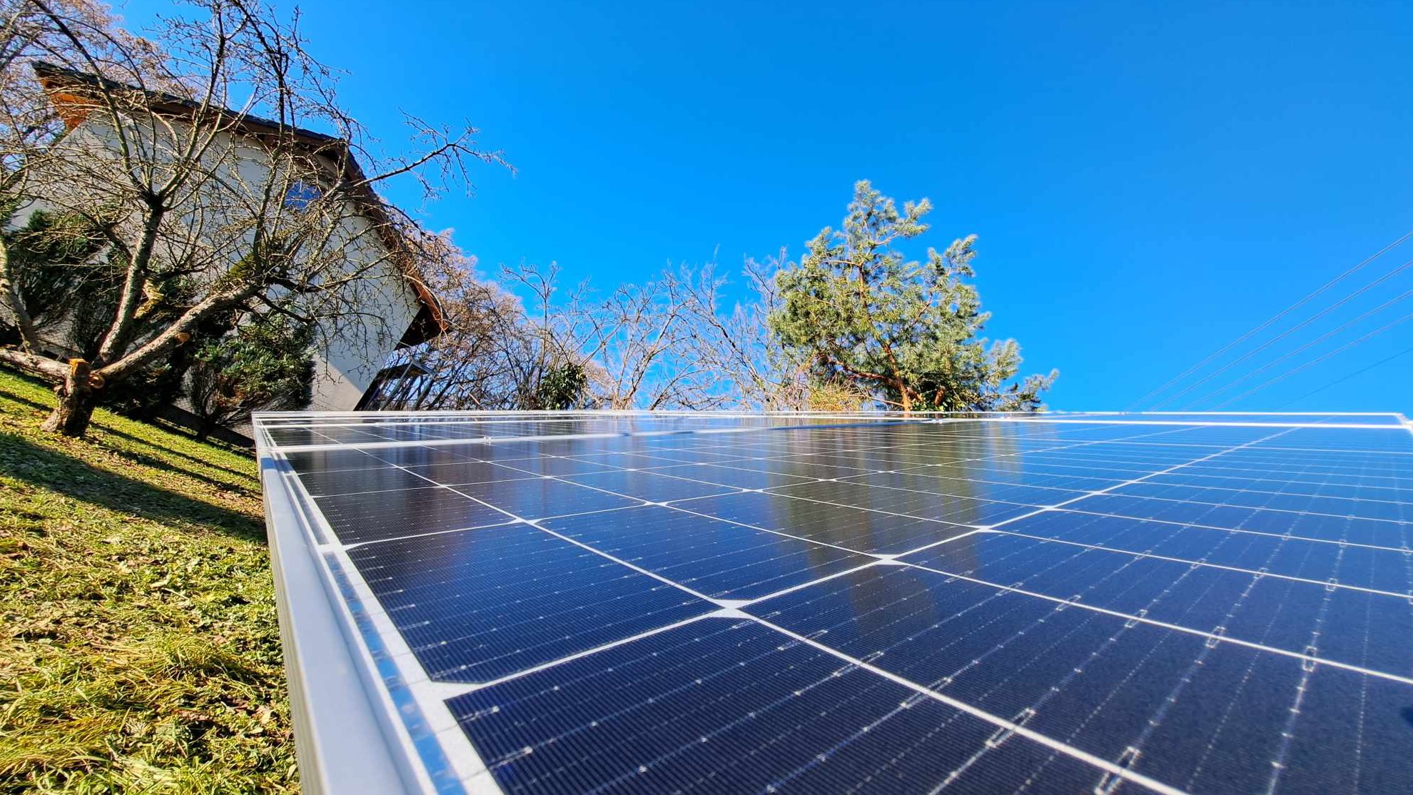 Nisko kątowe ujęcie paneli słonecznych na tle błękitnego nieba, z widocznym fragmentem domu i drzewami w tle.