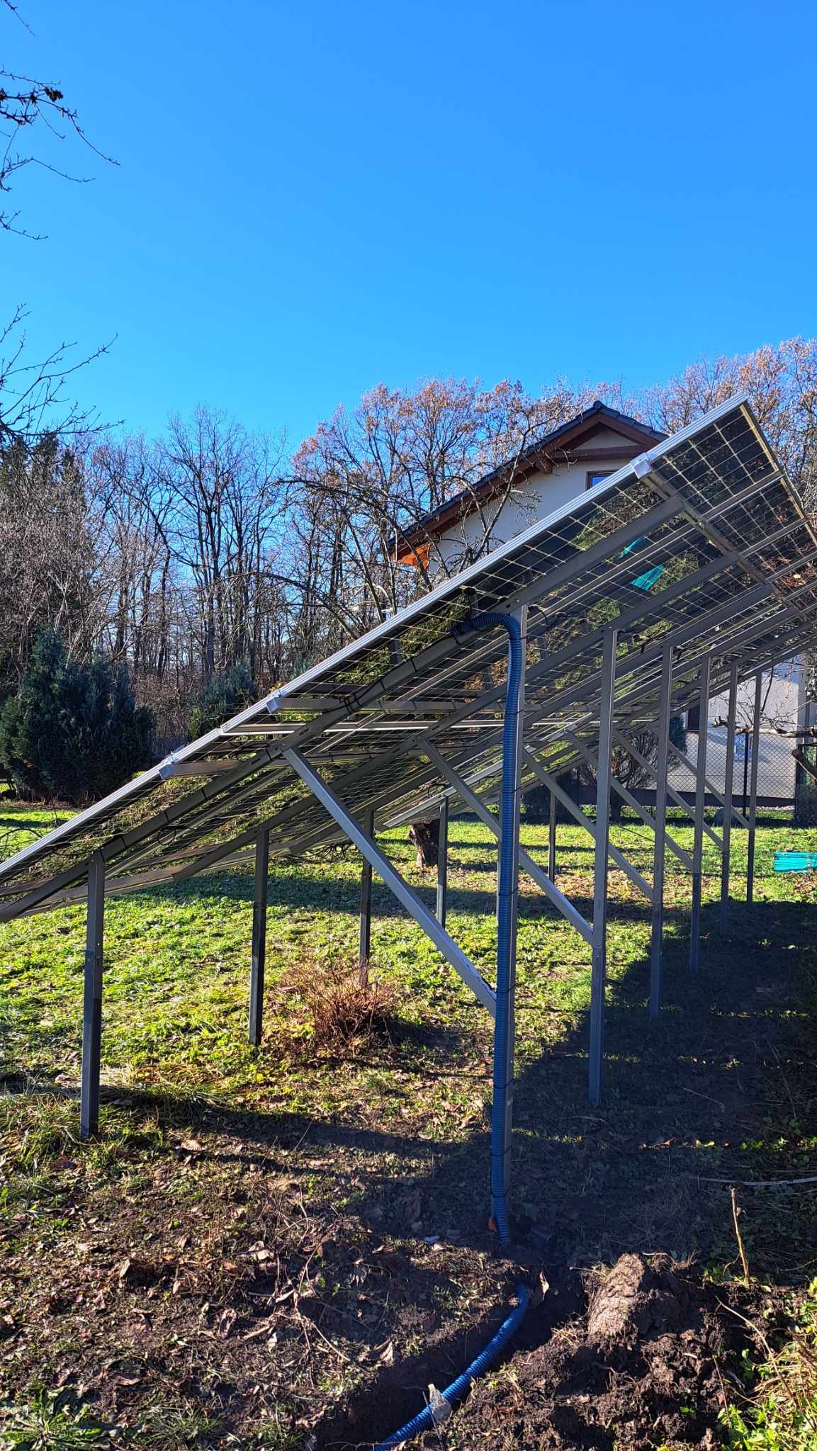 Konstrukcja wsporcza paneli słonecznych w trakcie montażu na zielonym trawniku, widoczne niebieskie rury i wykop w ziemi, dom w tle.