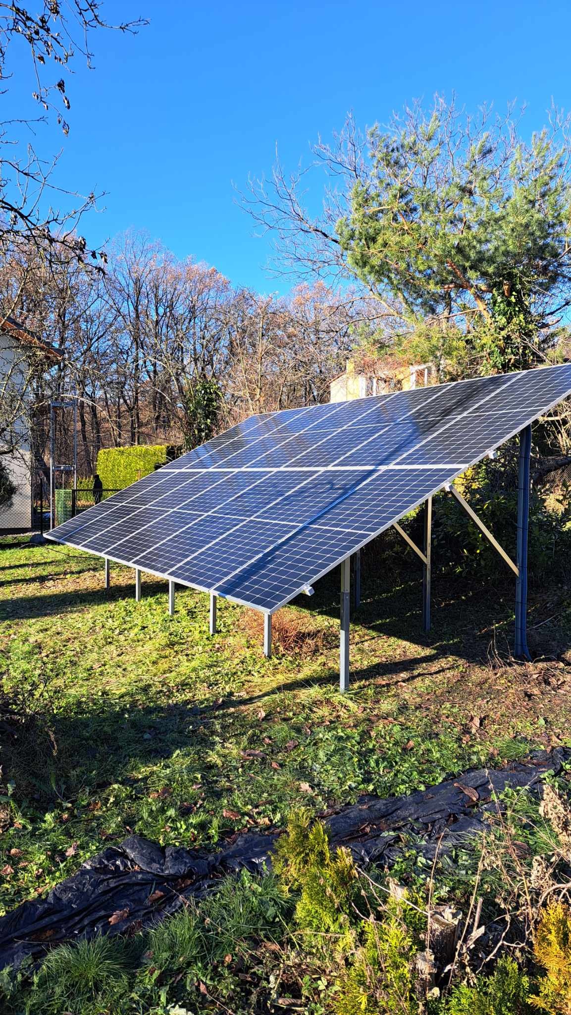 Panel fotowoltaiczny na metalowej konstrukcji, ustawiony na trawniku w ogrodzie, widoczny w słoneczny dzień, z drzewami w tle.