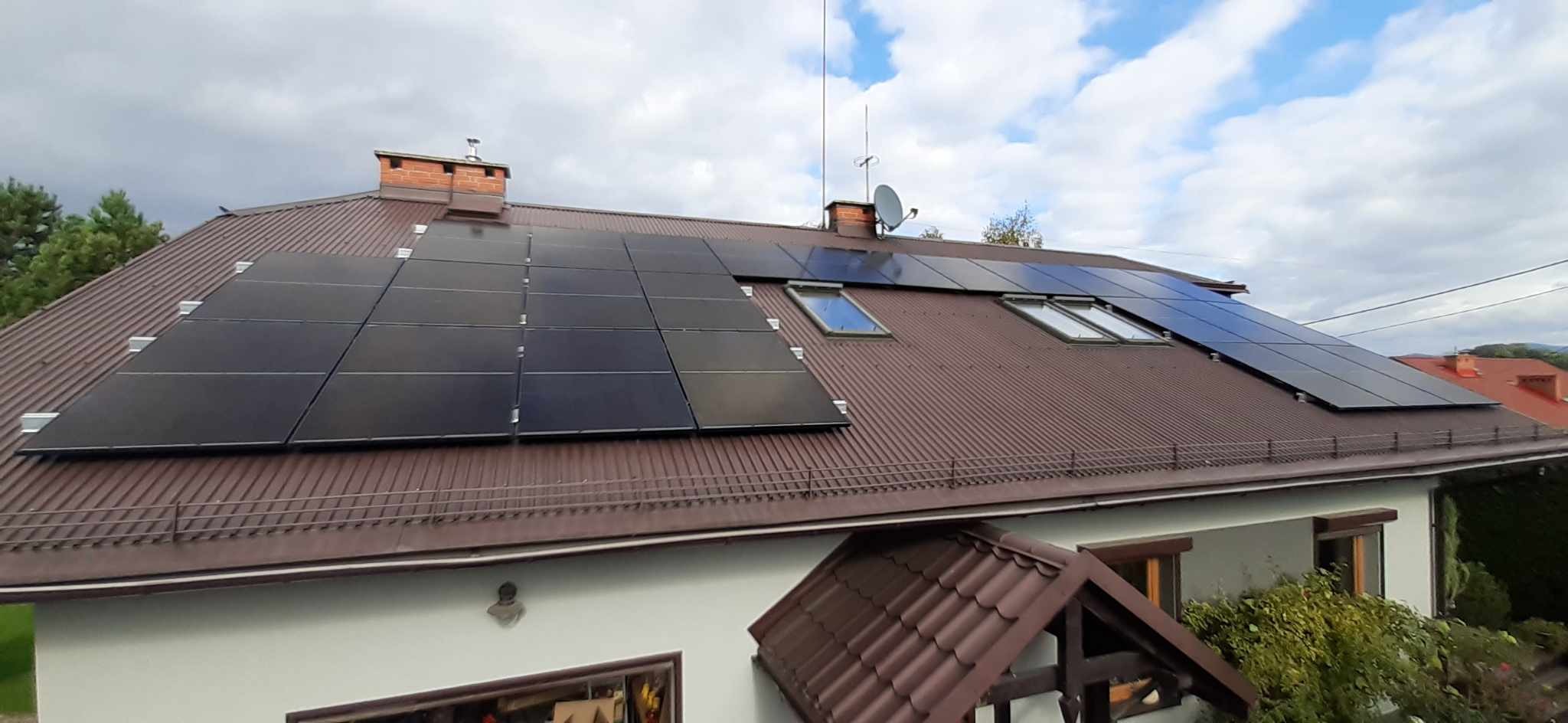 Brązowy dach dwuspadowy z panelami słonecznymi po lewej stronie i oknami dachowymi po prawej, widok z góry na dom z ogrodem.