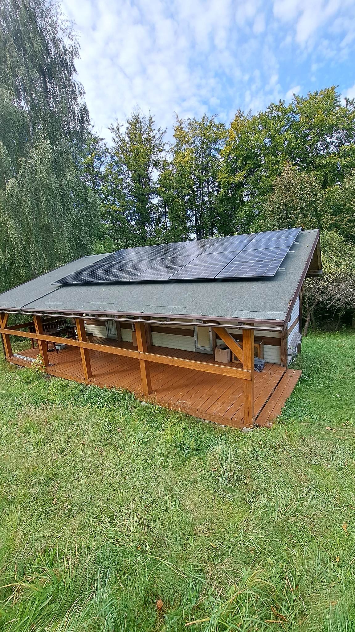 Dom z panelami słonecznymi na dachu, otoczony zielenią i drzewami, widok z dołu, lekko pochmurne niebo.