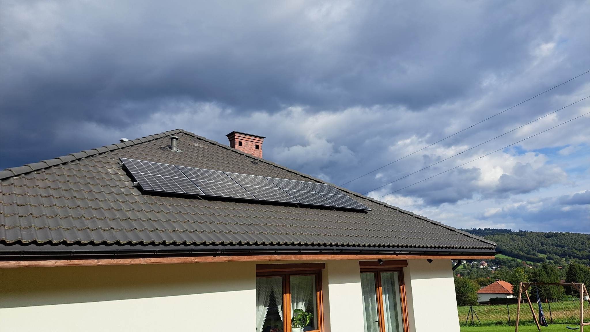 Panel słoneczny na dachu domu z ciemną dachówką, komin z cegły, pochmurne niebo w tle.