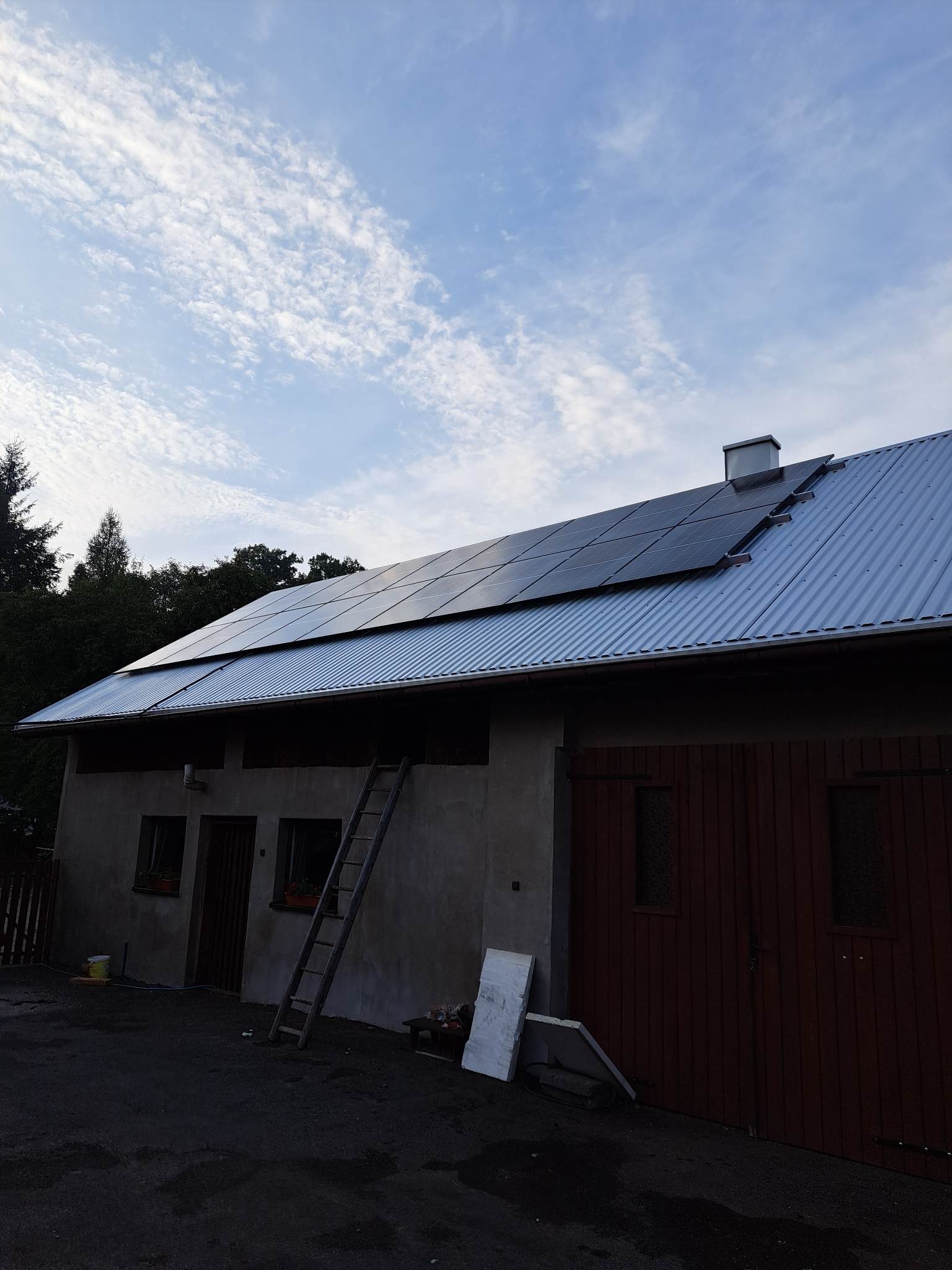 Instalacja paneli fotowoltaicznych na blaszanym dachu budynku gospodarczego; drabina oparta o ścianę, widoczne okna z kwiatami i drewniane wrota garażowe.