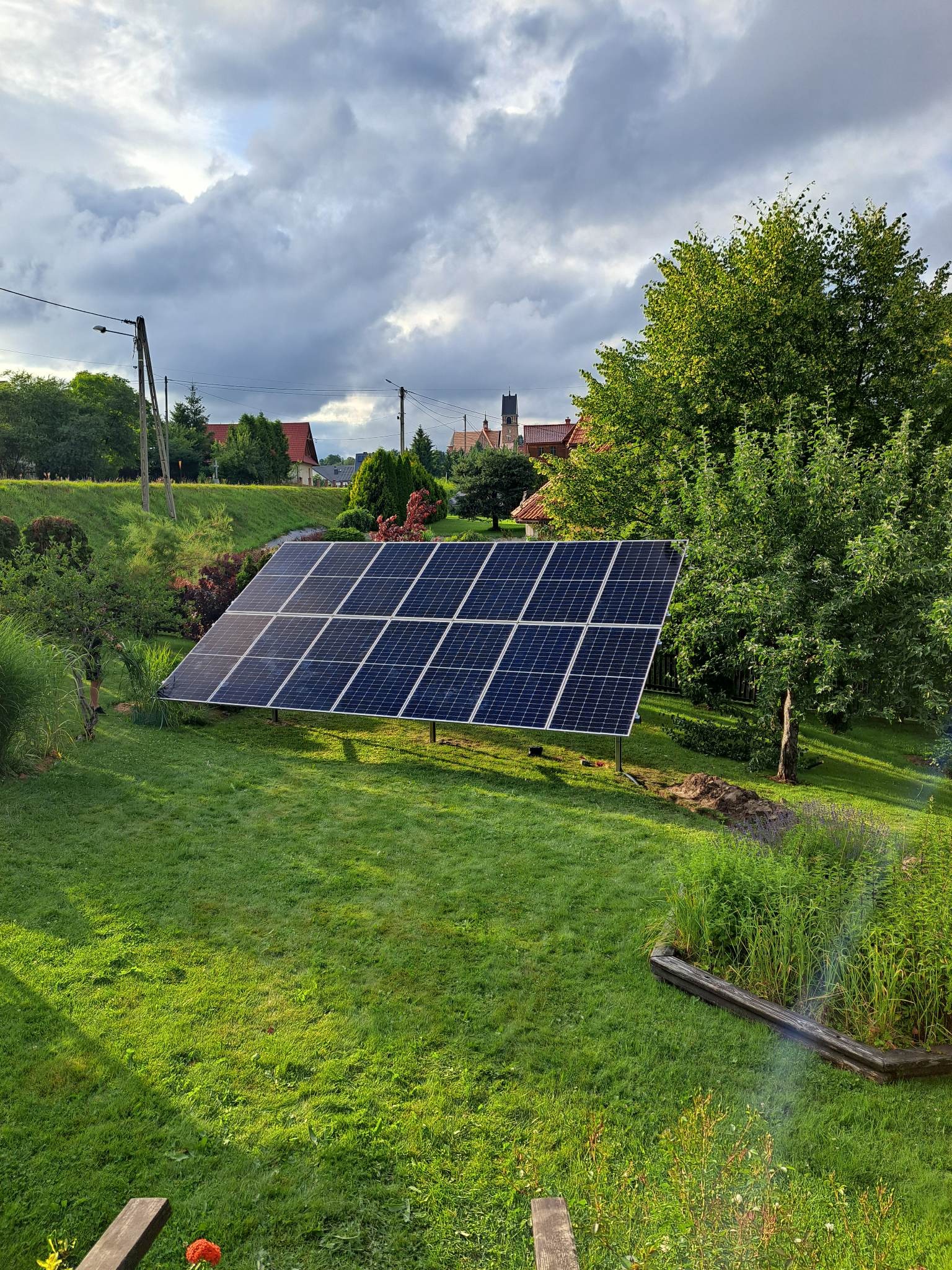 Panel słoneczny na metalowej konstrukcji w ogrodzie, w tle widoczne budynki mieszkalne i kościół pod pochmurnym niebem.