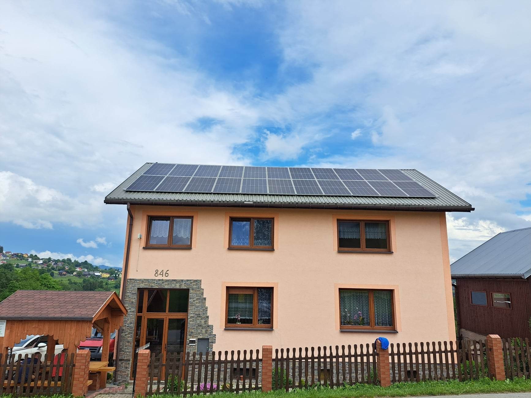 Dwupiętrowy dom z panelami słonecznymi na dachu, numer domu 846, widok z zewnątrz, drewniane ogrodzenie, w tle zielone wzgórza i błękitne niebo z chmurami.