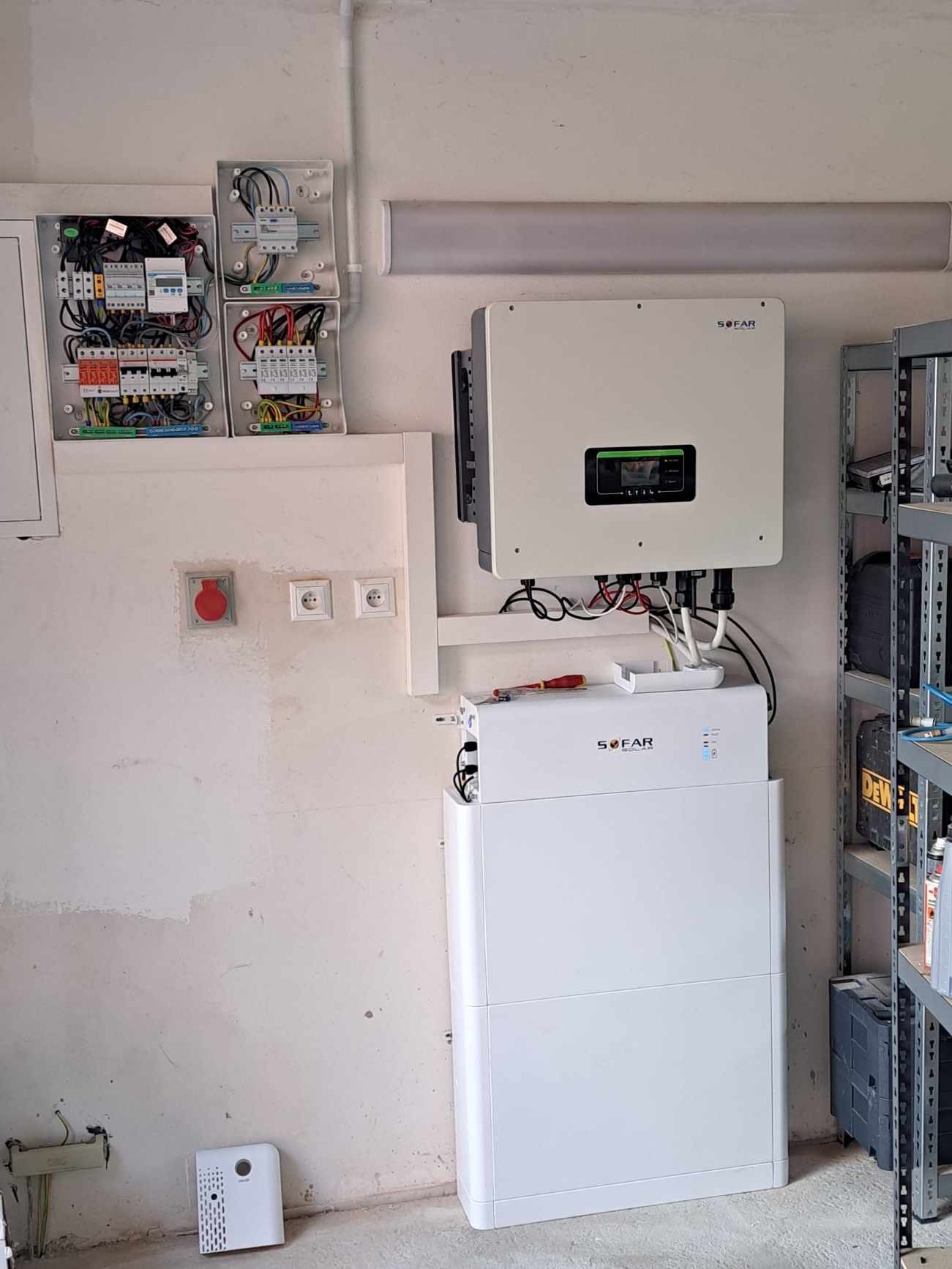 Biały falownik i akumulator energii SOFAR zamontowane na ścianie, widoczne otwarte skrzynki elektryczne z okablowaniem, czerwony wyłącznik bezpieczeństwa i gniazdka elektryczne na tle surowej ściany.