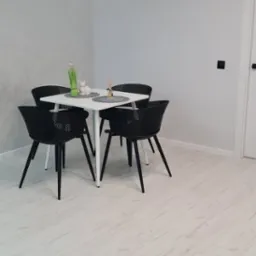 Jasne panele podłogowe w jadalni z białym stołem i czarnymi krzesłami, minimalistyczne dekoracje na stole, oświetlenie LED na suficie.