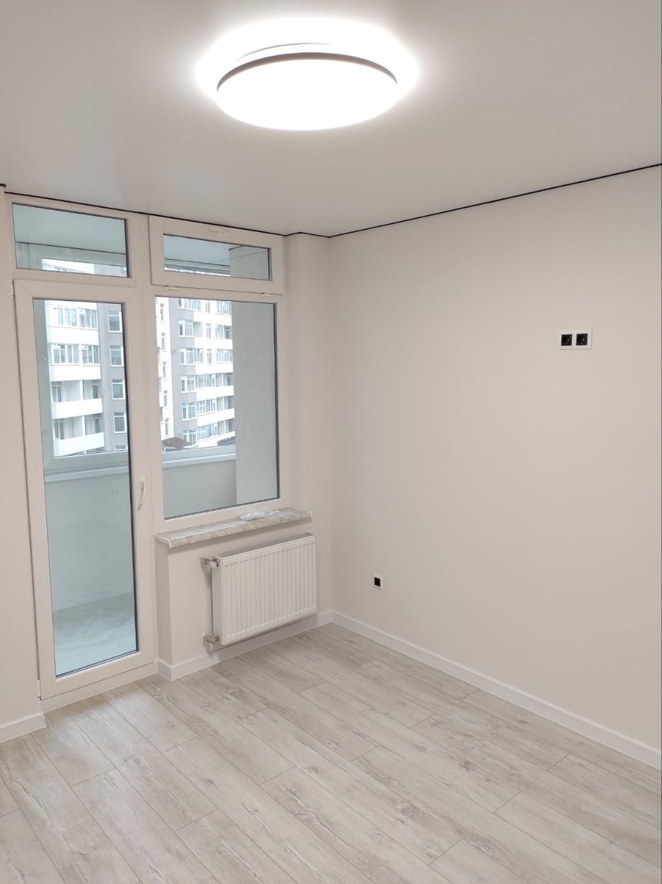 Jasne, puste pomieszczenie z panelami podłogowymi w odcieniach szarości, białymi ścianami z widocznymi gniazdkami elektrycznymi, grzejnikiem pod oknem balkonowym i okrągłą lampą sufitową.