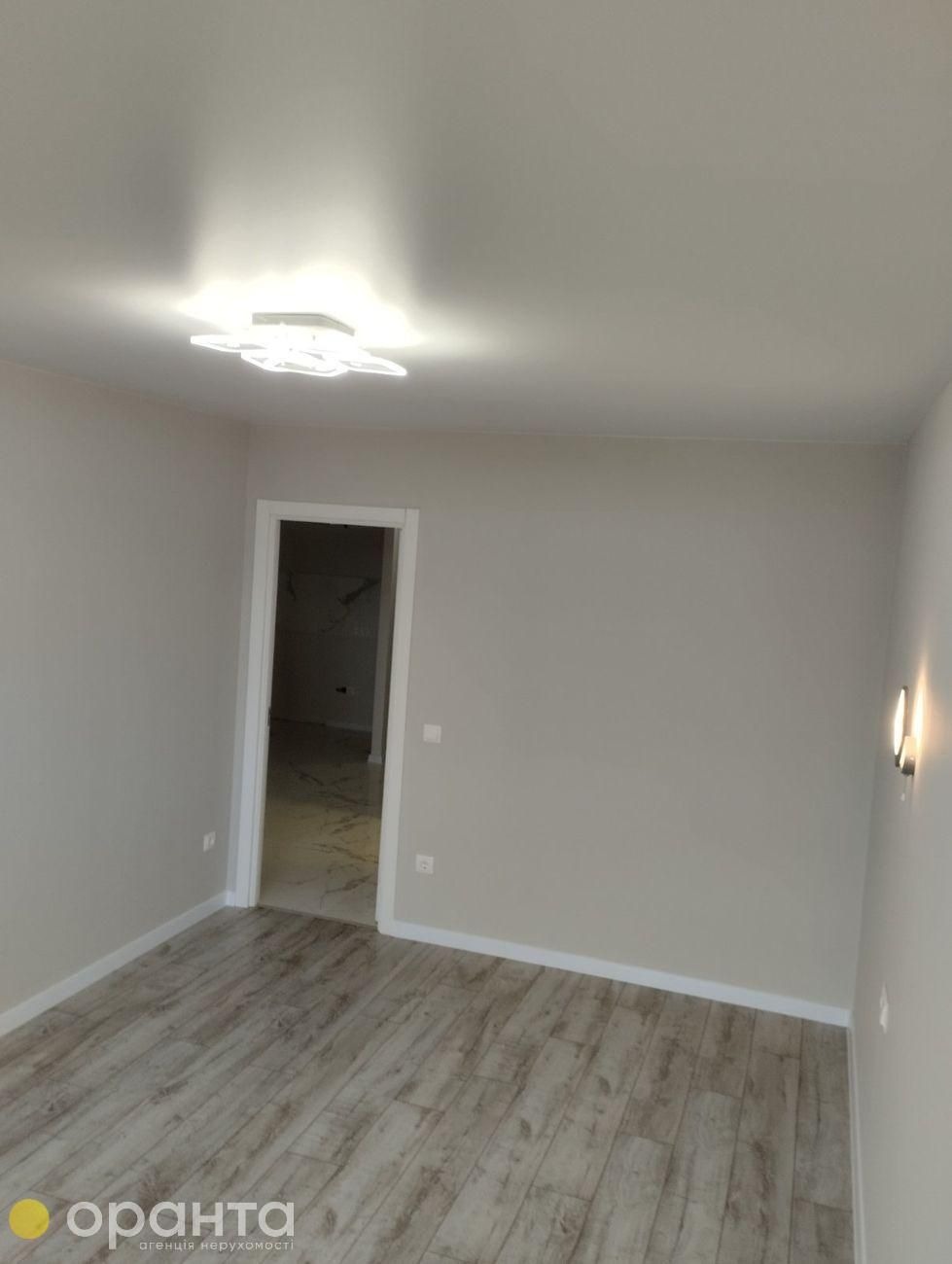 Jasne wnętrze pokoju po remoncie z panelami podłogowymi w odcieniach drewna, nowoczesnym oświetleniem sufitowym i widocznym fragmentem łazienki z marmurowymi płytkami przez otwarte drzwi.