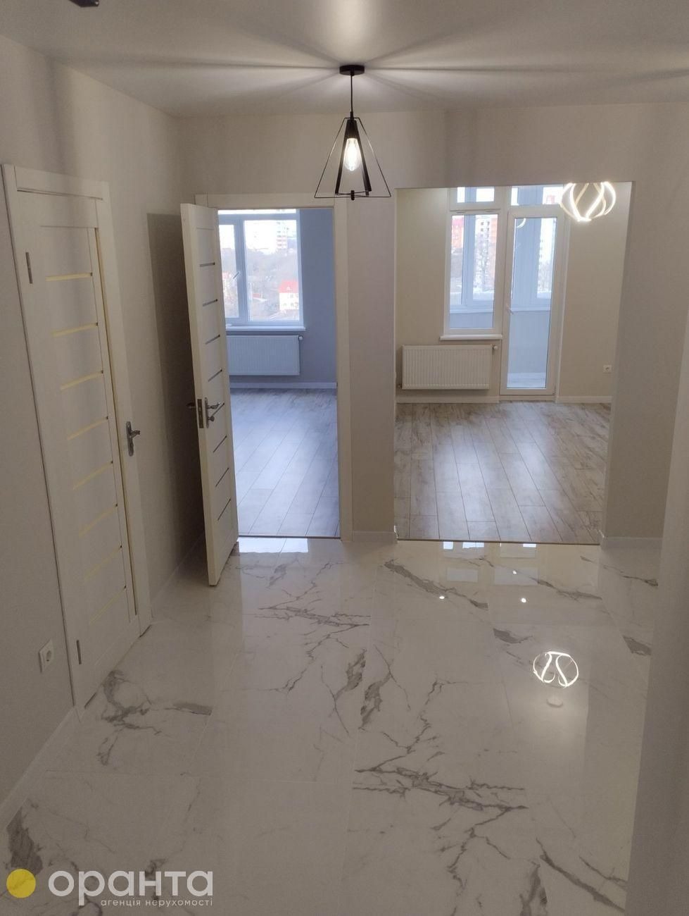 Wnętrze mieszkania po remoncie z białymi drzwiami, marmurową podłogą i widokiem na pokoje z drewnianą podłogą, oświetlone nowoczesną lampą.