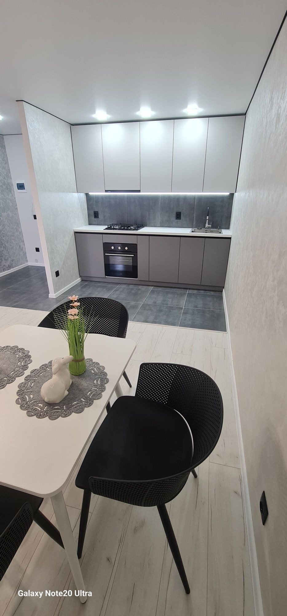 Nowoczesna kuchnia w odcieniach szarości i bieli, z minimalistycznymi szafkami, płytą grzewczą i piekarnikiem. Na pierwszym planie biały stół z dekoracją: ceramiczny zajączek i bukiet różowych...