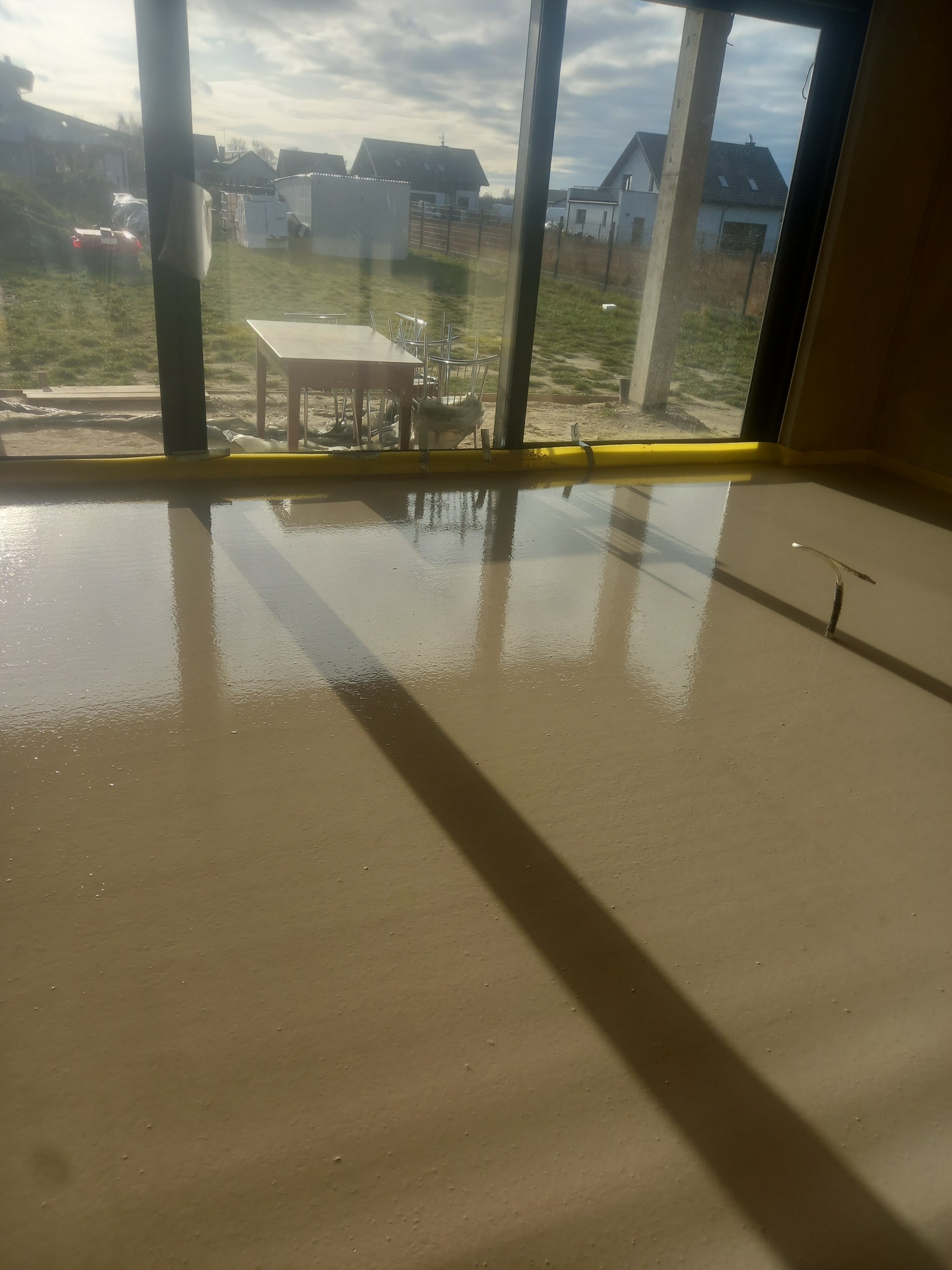 Świeżo wylana, gładka posadzka w jasnym odcieniu beżu, z widocznymi refleksami światła słonecznego padającego przez duże okno tarasowe, żółta taśma piankowa przy oknie.