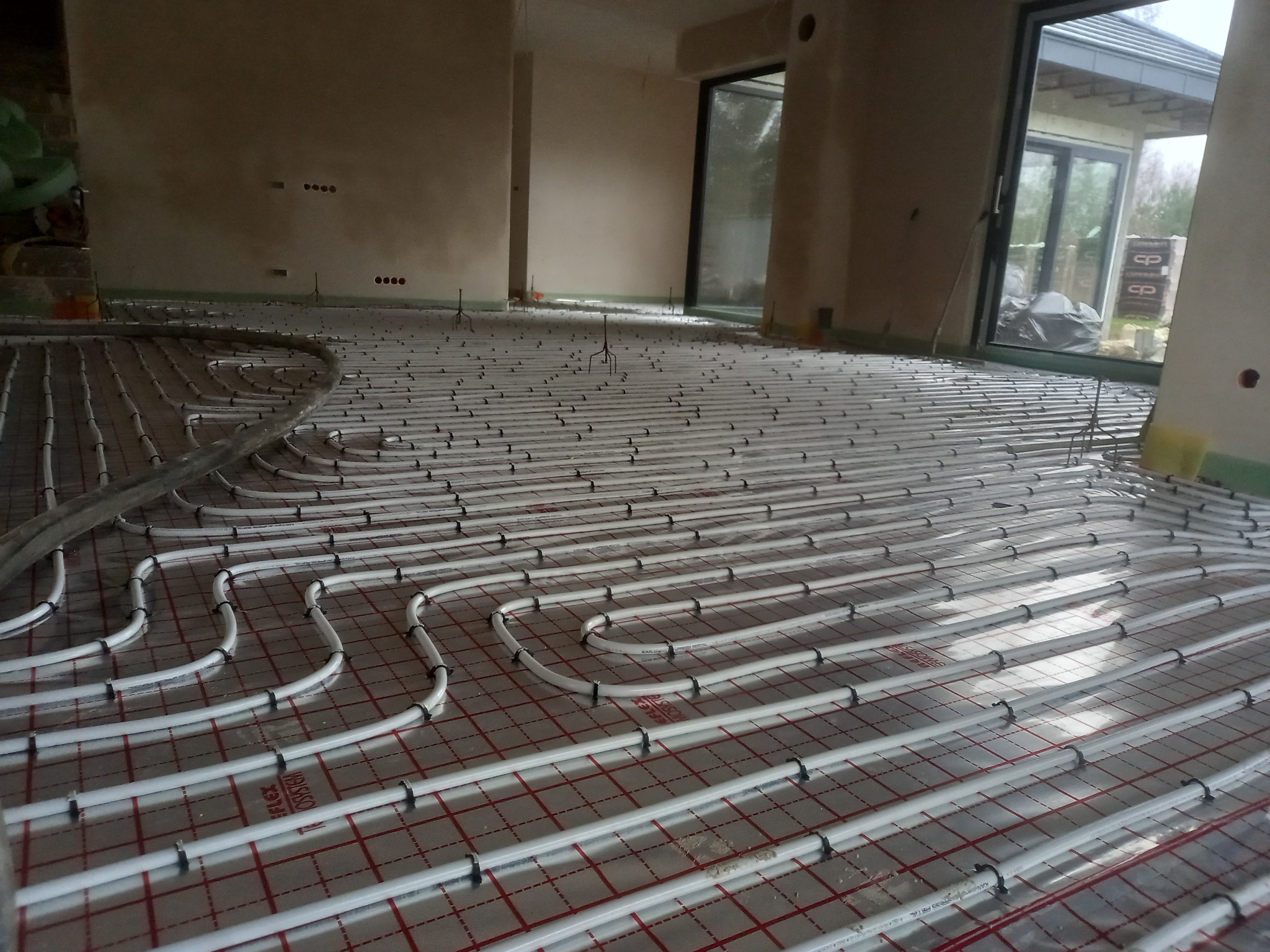 Rozłożone białe rury ogrzewania podłogowego na folii z czerwoną siatką w surowym wnętrzu z dużymi oknami.